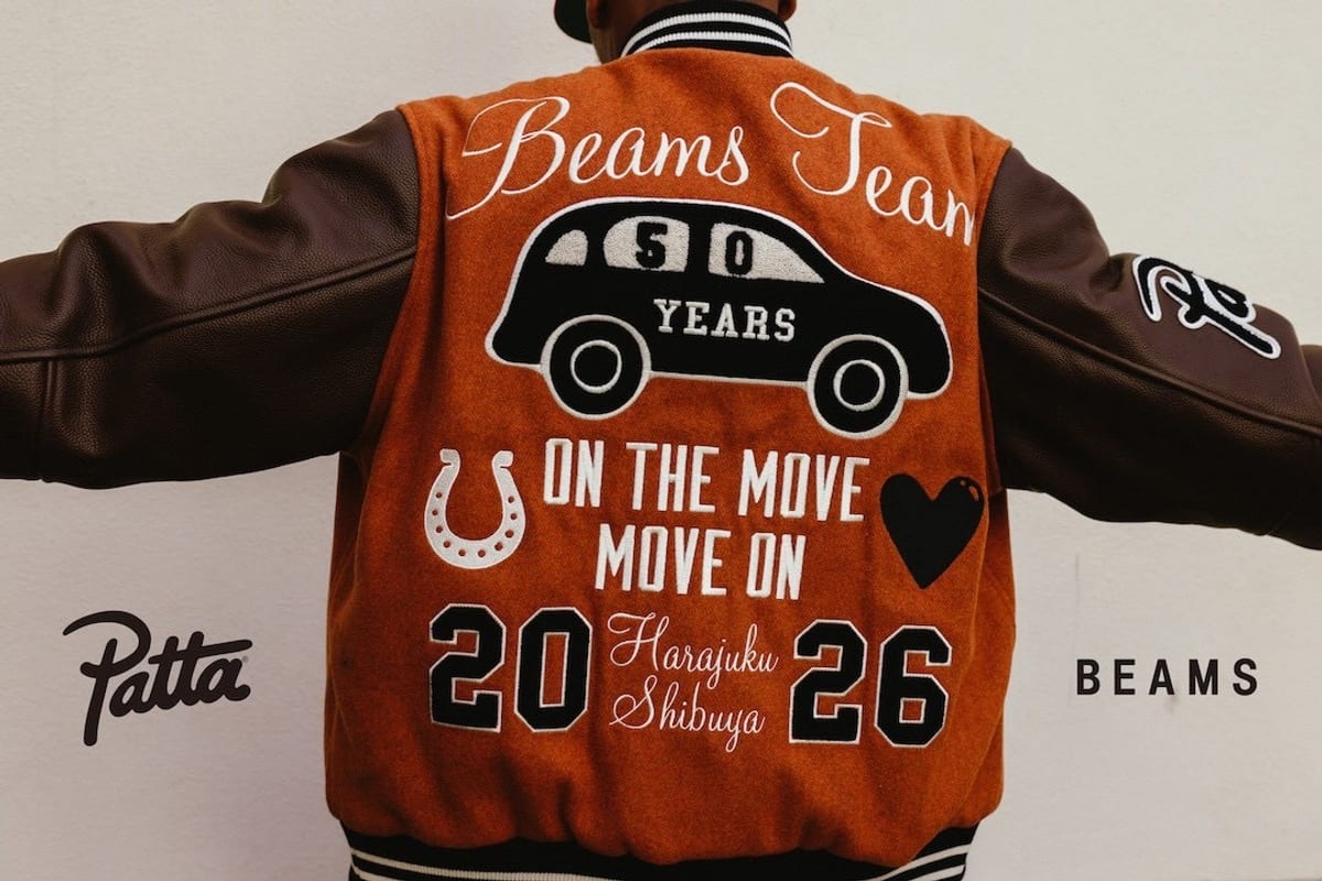 The back of the rust-orange varsity jacket worn with arms outstretched against a white wall: 'Beams Team' in white script, a black car marked '50 Years', a horseshoe, 'On the move, move on', a black heart, and '20 26' around 'Harajuku Shibuya', with the Patta and BEAMS logos set either side of the photo