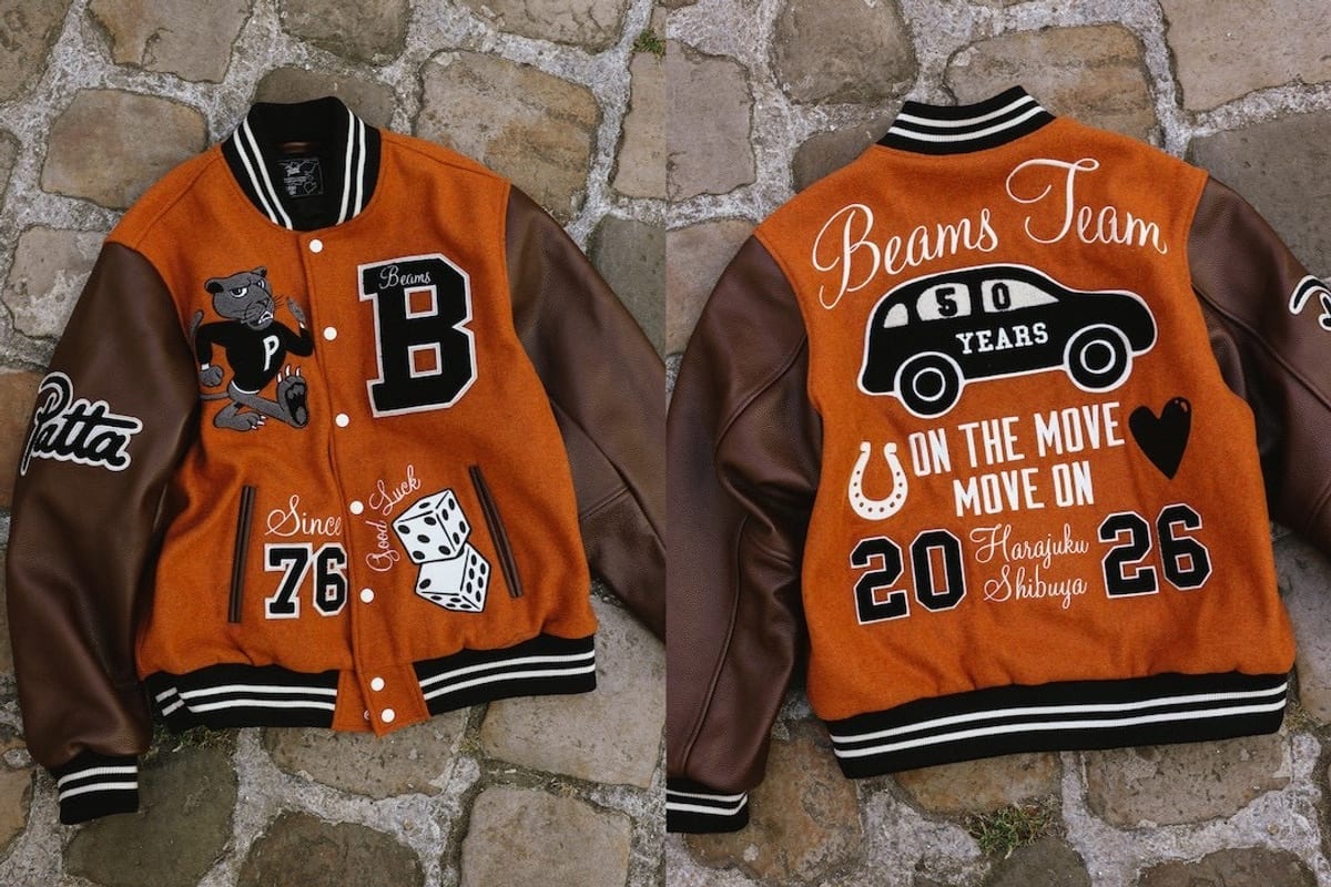 The front and back of a rust-orange wool varsity jacket with brown leather sleeves laid on cobblestones: the front has a cartoon panther with a P, a black 'B' with 'Beams', 'Since 76' and a pair of dice reading 'Good Luck', a Patta script on the sleeve; the back reads 'Beams Team' over a car marked '50 Years', 'On the move, move on' and '2026, Harajuku Shibuya'