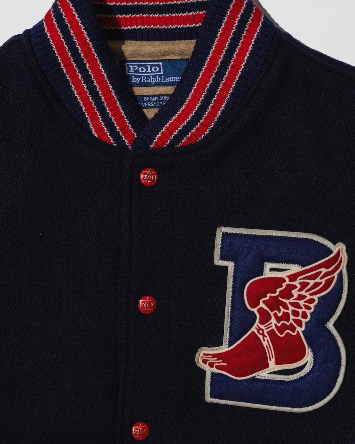 Close-up of the B-WING patch on the Polo Ralph Lauren for BEAMS letterman jacket: a red winged foot over a navy chenille B, below a navy, red and white striped collar