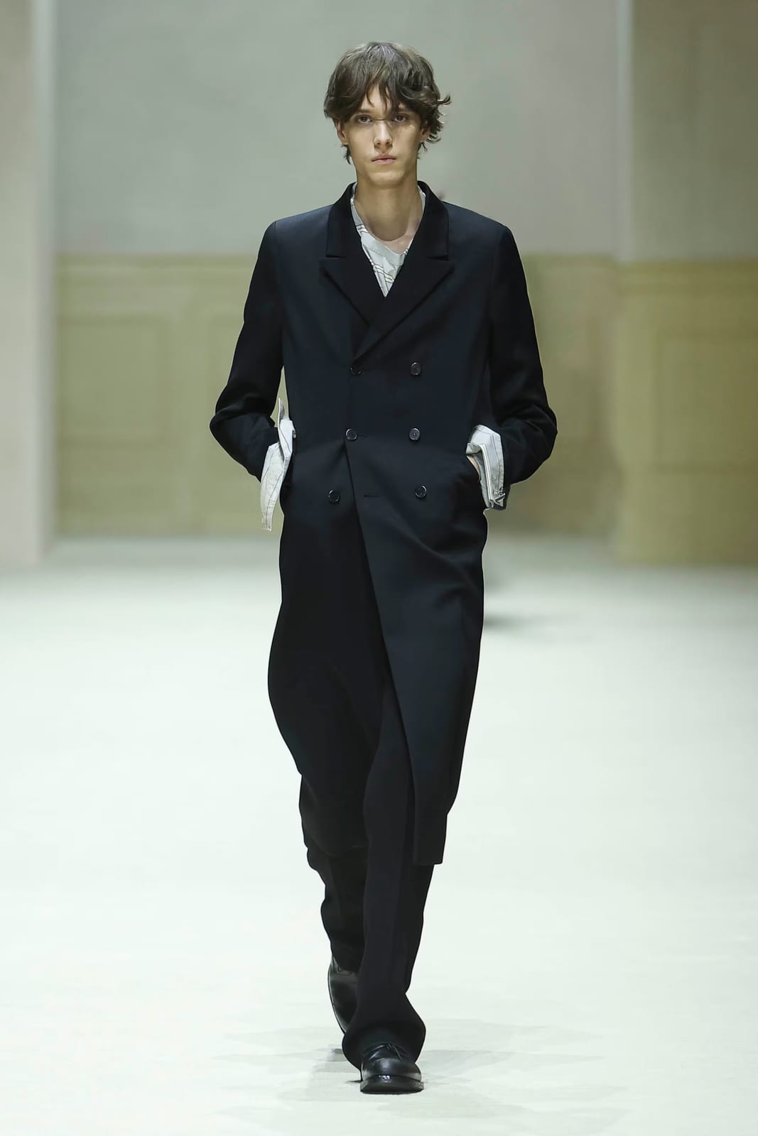 The opening look of Prada's Fall/Winter 2026 menswear show: a long black double-breasted topcoat buttoned high, with white shirt cuffs showing at the pockets