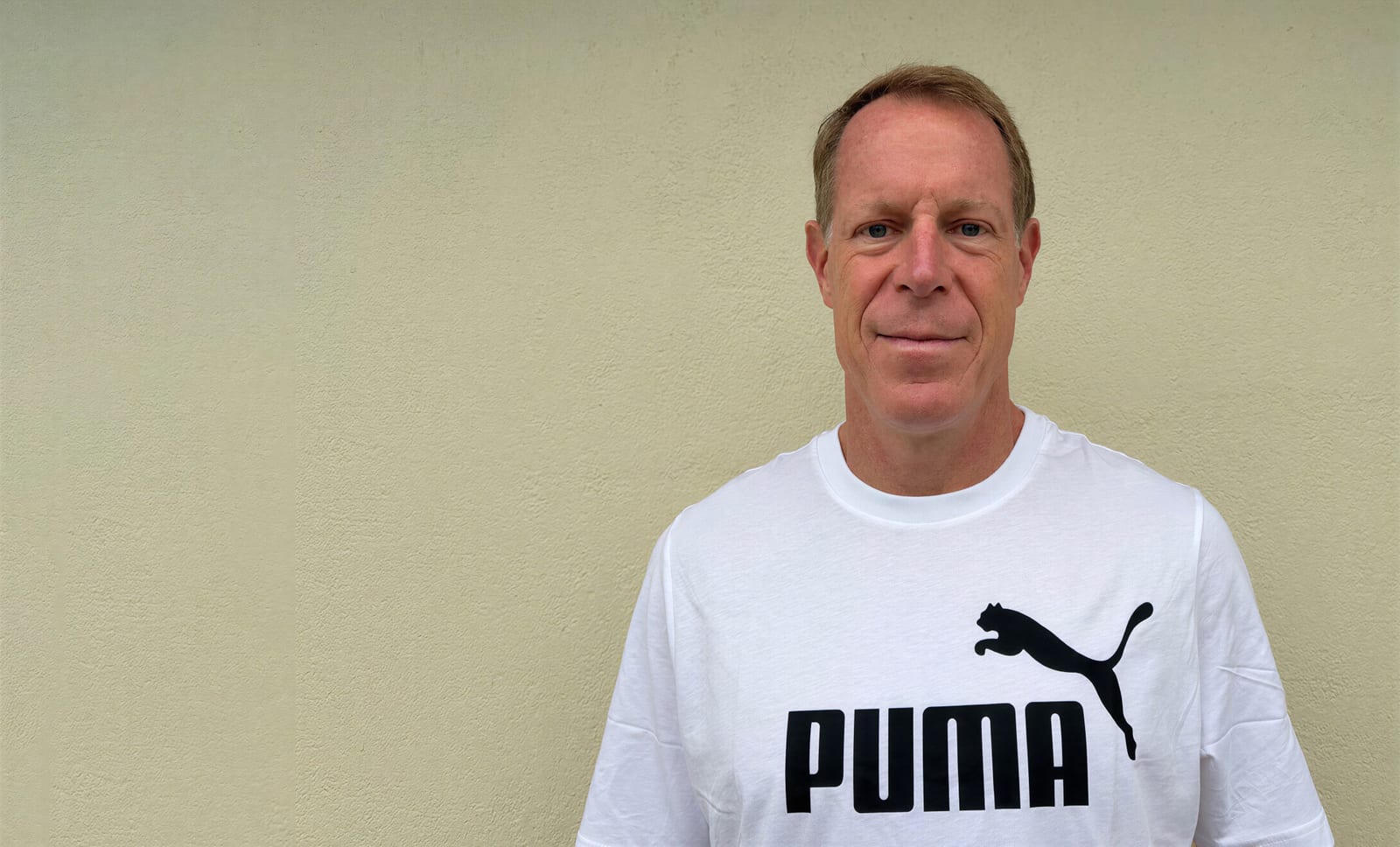 Arne Arens, a man with short light brown hair, in a white T-shirt with the black PUMA logo and leaping cat, standing against a pale beige wall
