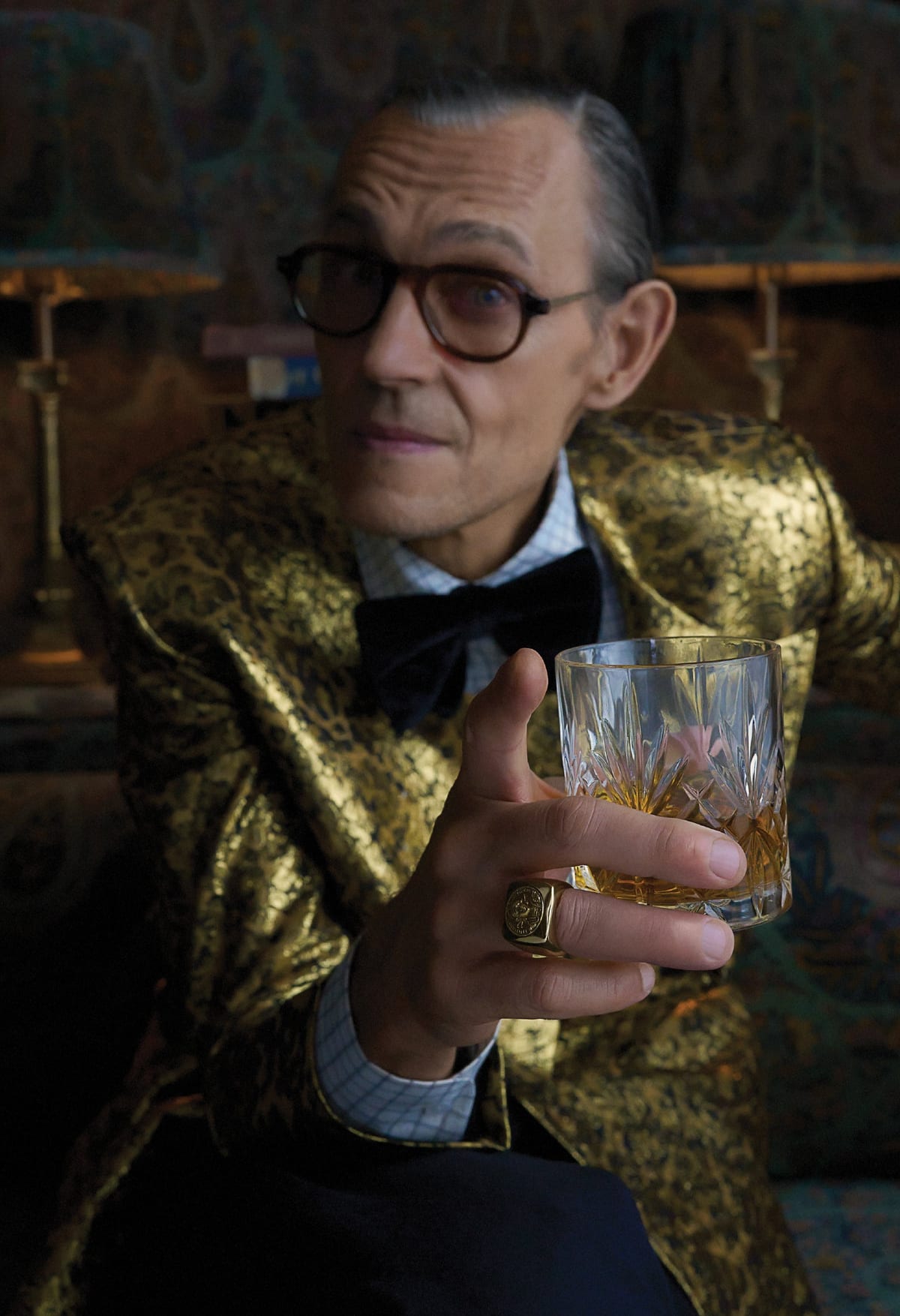 A man in a gold brocade jacket and bow tie wearing a chunky gold signet ring from the collaboration, holding a crystal glass