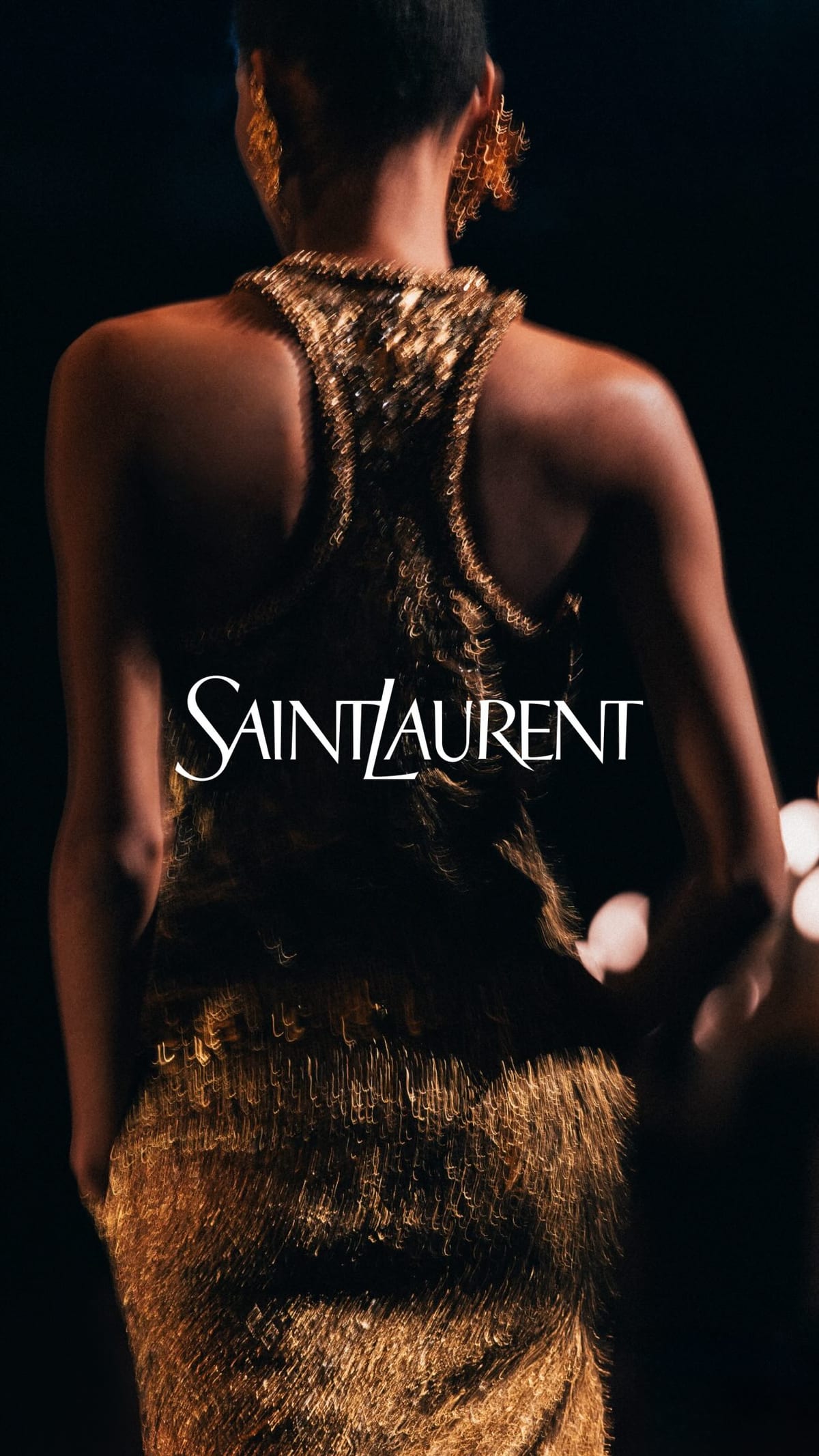 A model seen from behind, walking away in a gold fringed dress at the finale of Anthony Vaccarello's Spring 2027 show for Saint Laurent, his last for the house, with the Saint Laurent logo across the image