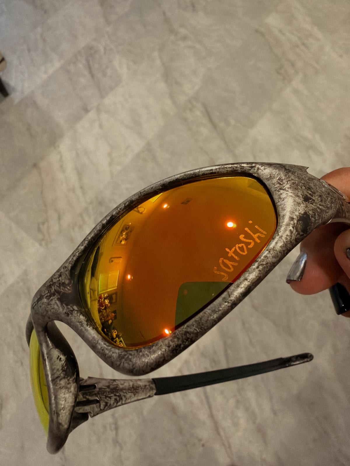 Close-up of the Satoshi Nakamoto x Oakley sunglasses: a distressed silver wraparound frame with an orange mirrored lens marked satoshi, held over a marble floor, from Jaysse Lopez's Instagram post