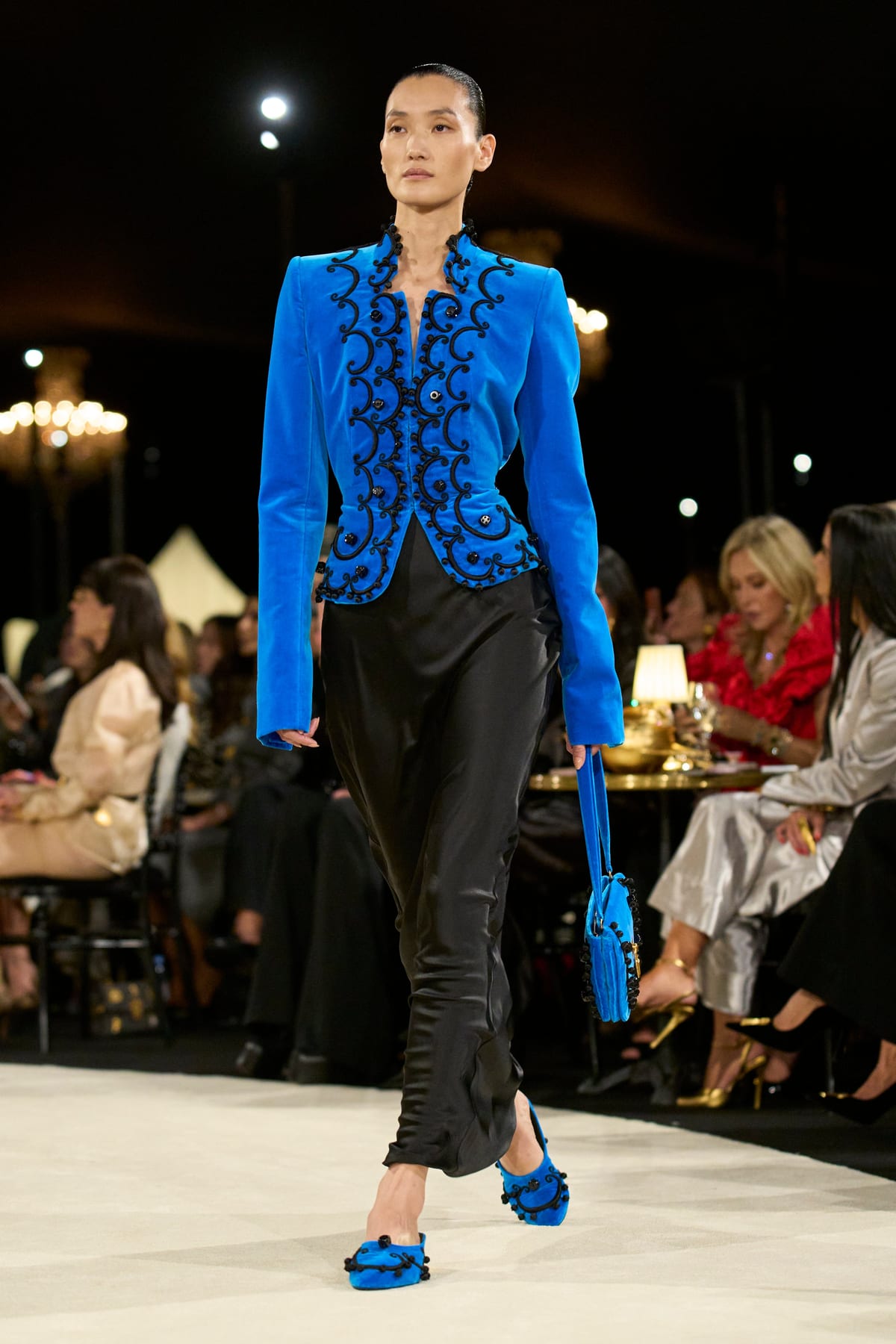 A model in a kingfisher blue jacket with black braided trim, over black satin trousers and blue shoes