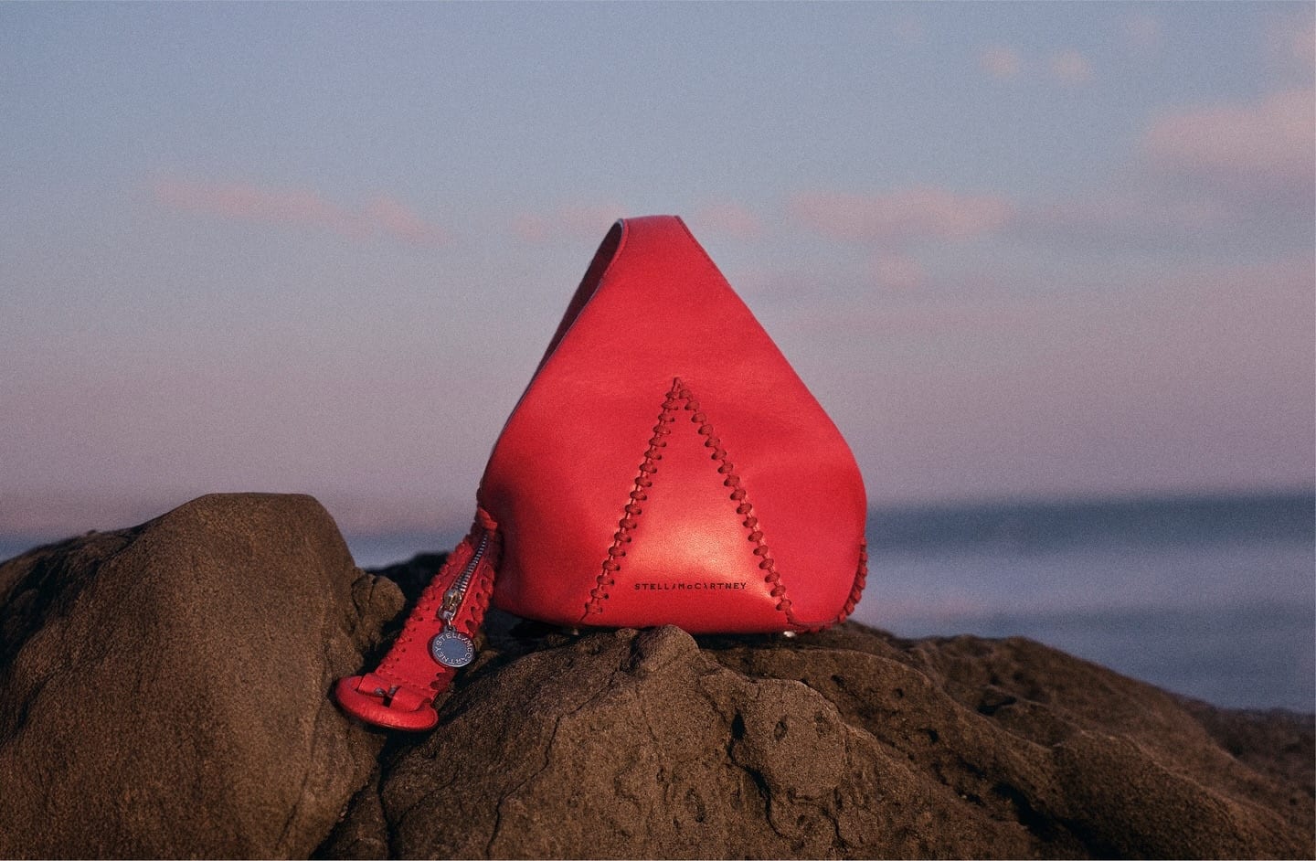 A red Stella McCartney Appaloosa wristlet with a pointed, teardrop shape, a triangular panel edged in whipstitching and a logo zip pull, set on a dark rock by the sea at dusk