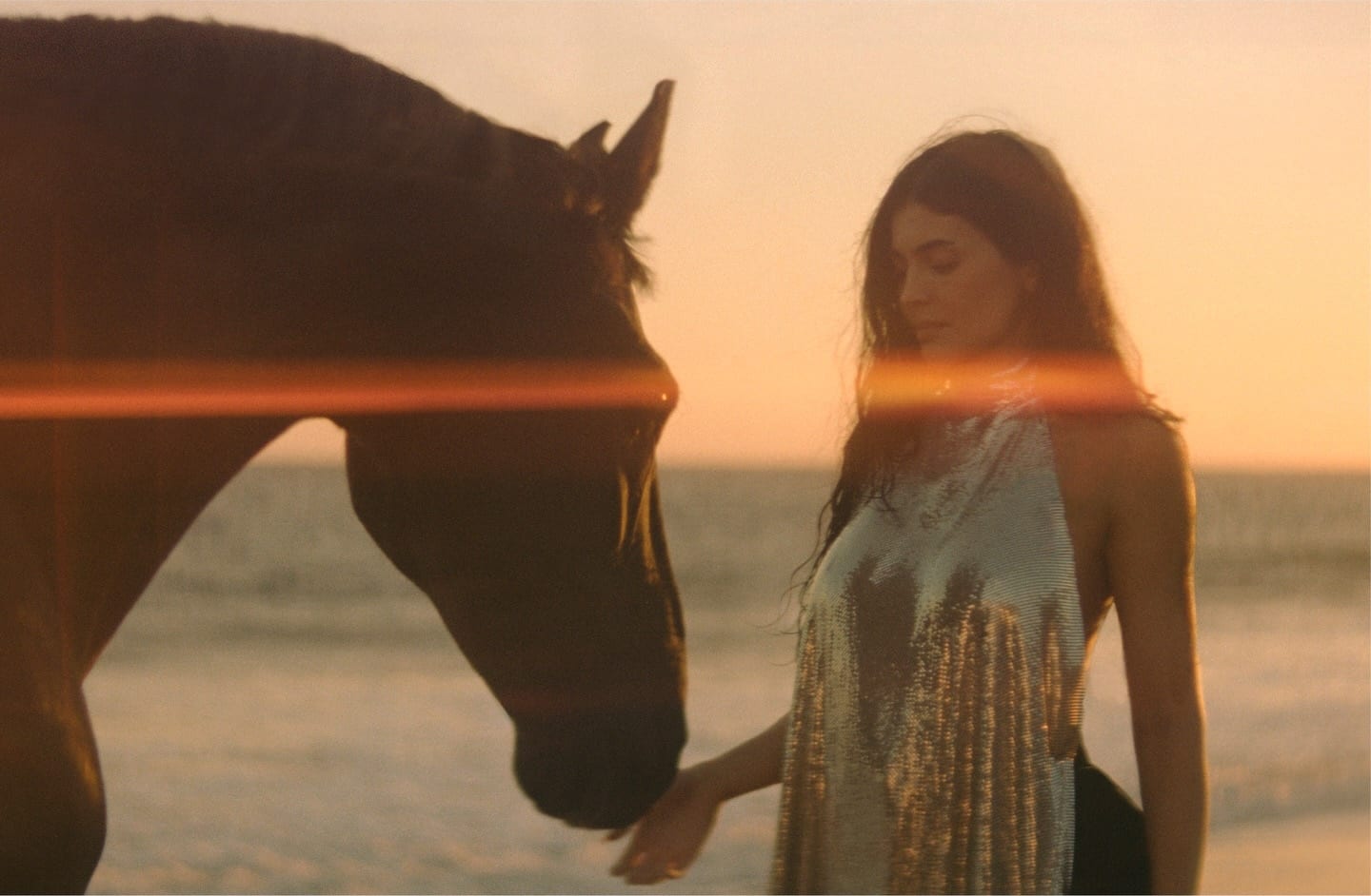 Kylie Jenner in a silver chainmail halterneck top stands on a beach at sunset, her hand held out under the muzzle of a dark horse, with an orange lens flare across the frame