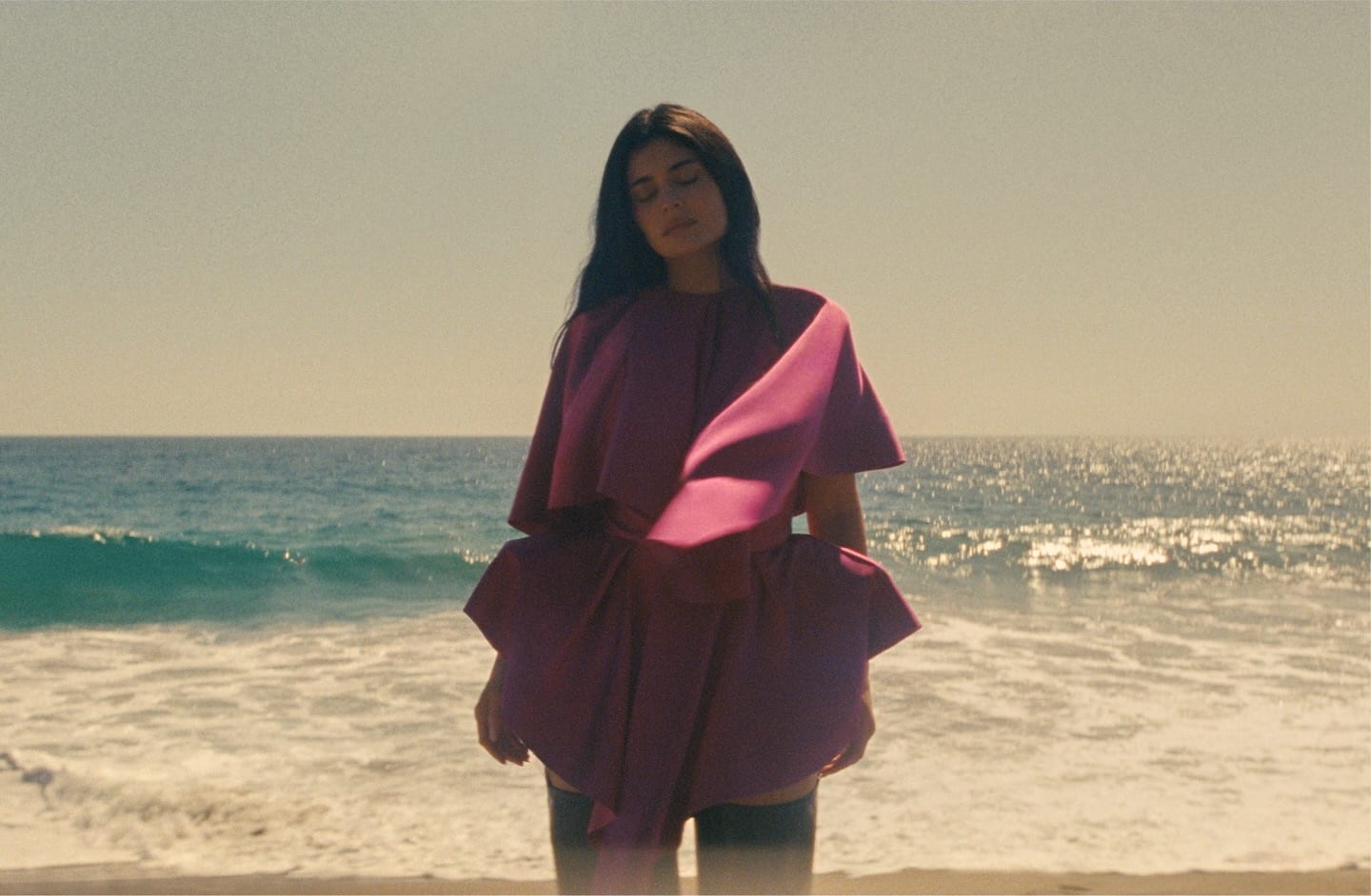 Kylie Jenner stands with her eyes closed at the edge of the sea in a magenta layered satin mini dress with a draped cape-like top and sculpted peplum, over dark over-the-knee boots