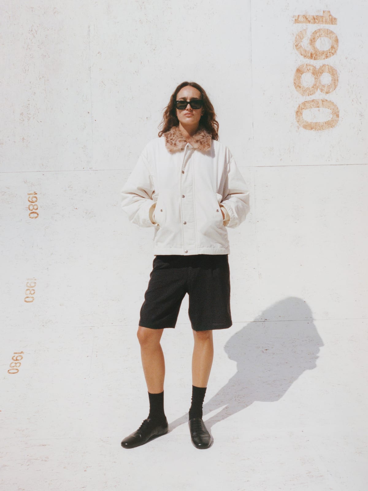 A woman in sunglasses stands in front of a white wall stencilled with 1980 in a cream snap-front jacket with a leopard-print collar, black shorts, black socks and black lace-up shoes