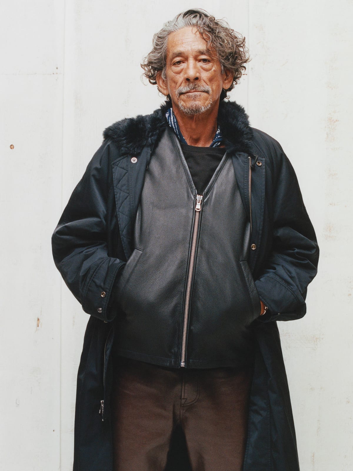 A grey-haired man with a moustache stands against a white wall in a black leather zip jacket under a long dark coat with a fur collar, hands in his pockets