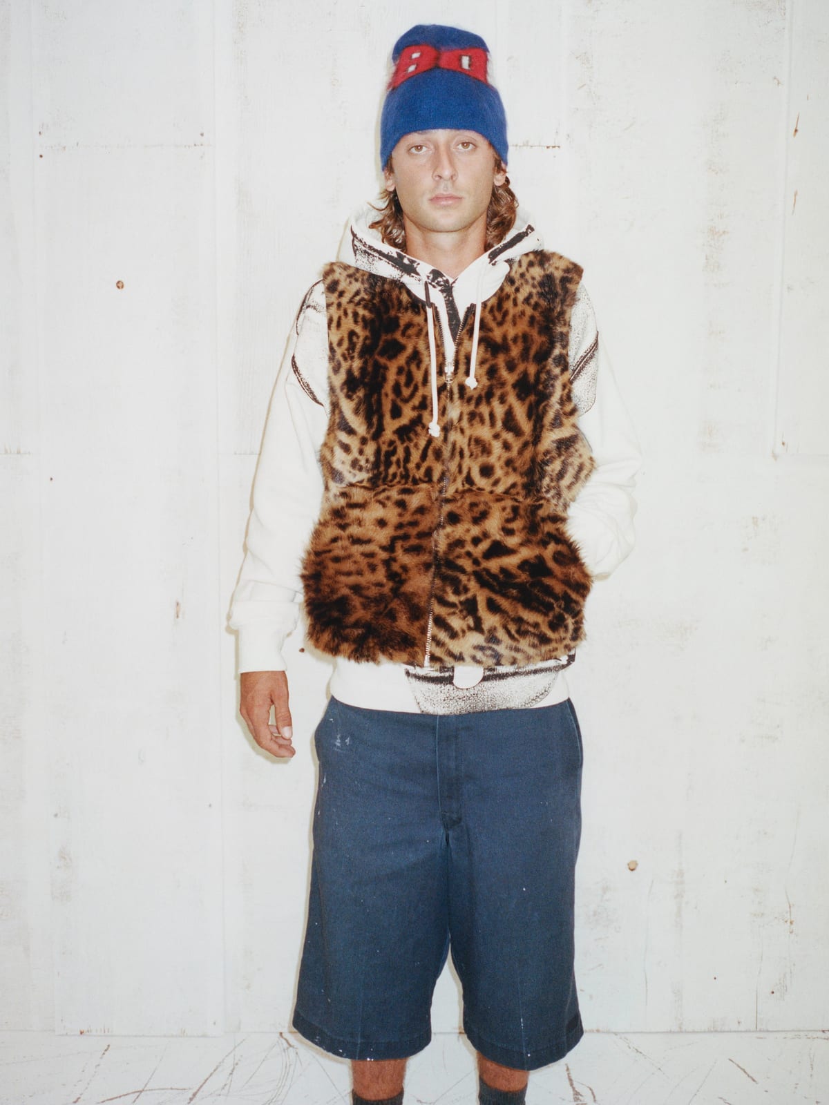 A young man in a blue beanie wears a leopard-print pile vest over a white hoodie with navy work shorts, against a white wall