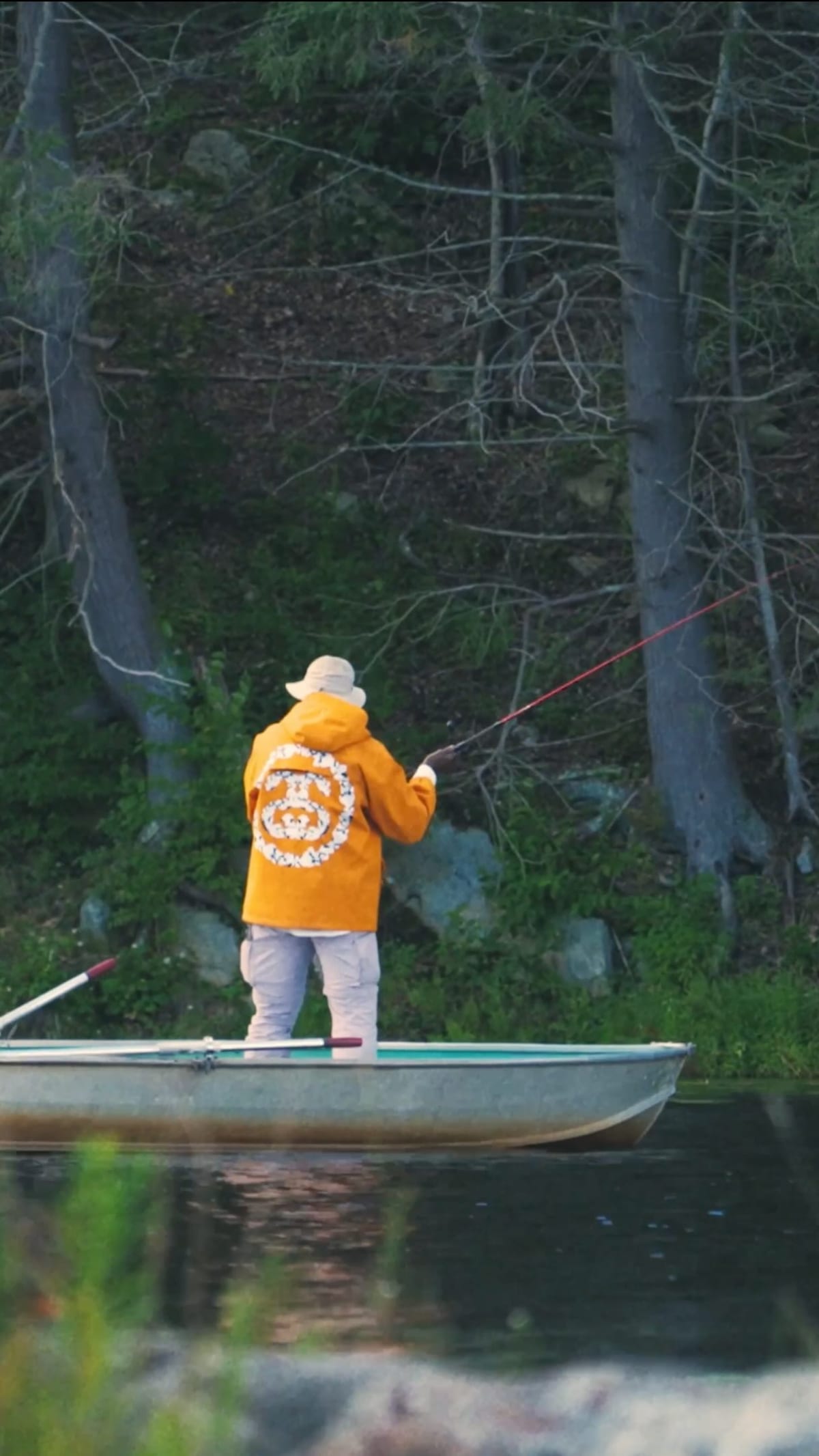 A man in a bucket hat and an orange hooded jacket stands in an aluminium rowboat on a wooded lake, casting a fishing rod, with a ring of white flowers around a Stüssy circle logo on the jacket's back