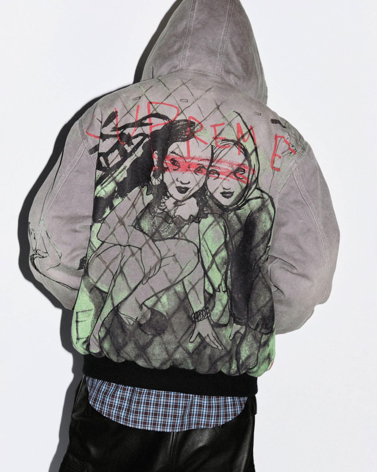The back of the Supreme/Rita Ackermann Hooded Work Jacket in washed grey canvas, printed with Rita Ackermann's drawing of two faces over a chain-link fence in black and green, with SUPREME scrawled in red, from Supreme's Fall/Winter 2026 lookbook