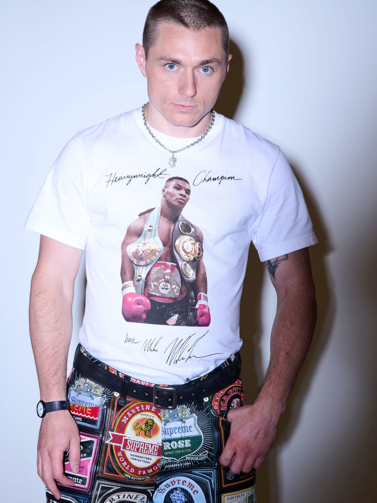 A model in a white T-shirt printed with a photo of a boxer wearing three championship belts and red gloves, with Heavyweight Champion written above in handwriting and a signature below, worn with the patchwork leather jeans