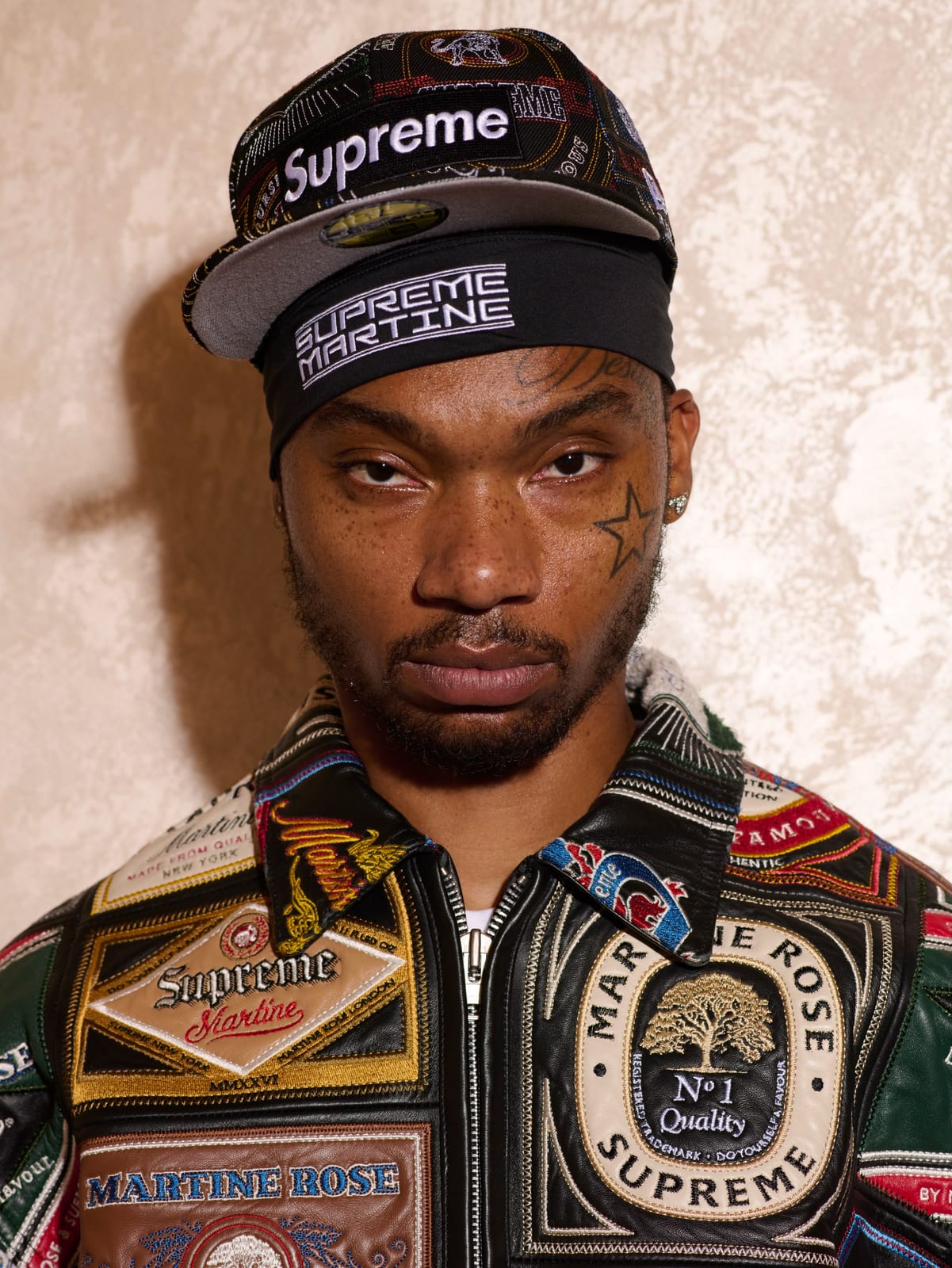 A model in a black Supreme New Era cap and a black Supreme Martine headband, wearing a black leather zip jacket patched all over with embroidered Martine Rose and Supreme crests, in red, green, cream and gold