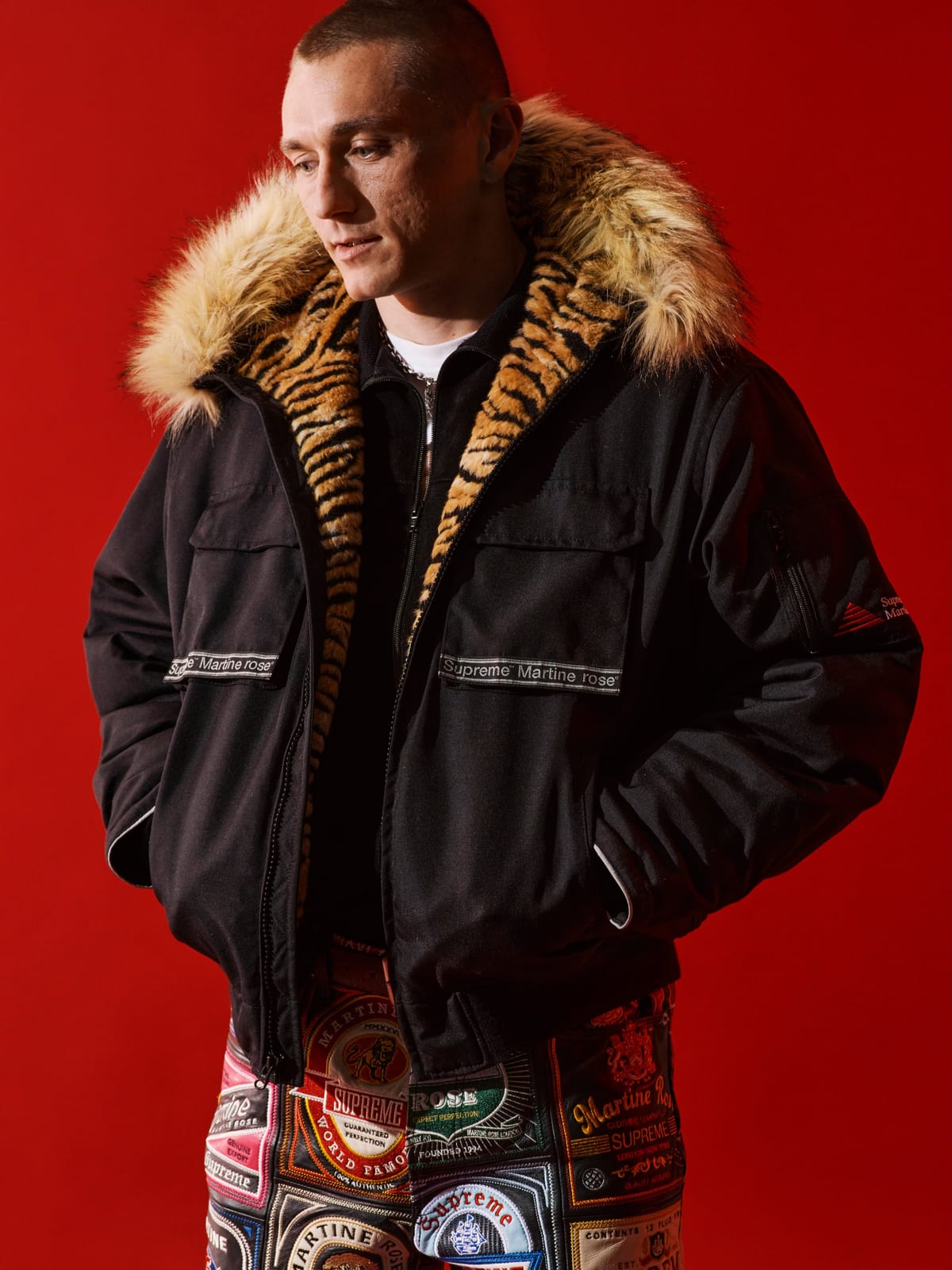 A model in a black Supreme/Martine Rose hooded jacket with a faux fur trim and a tiger-stripe lining, standing against a red backdrop in the patchwork leather jeans