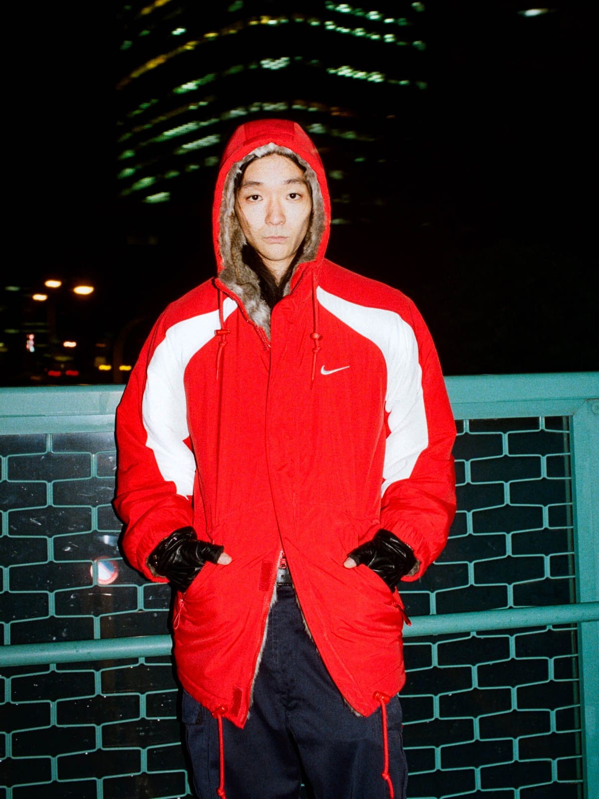 The Supreme x Nike Faux Fur Reversible Parka worn on its nylon side, red with white panels and a Nike Swoosh on the chest, on a city footbridge at night