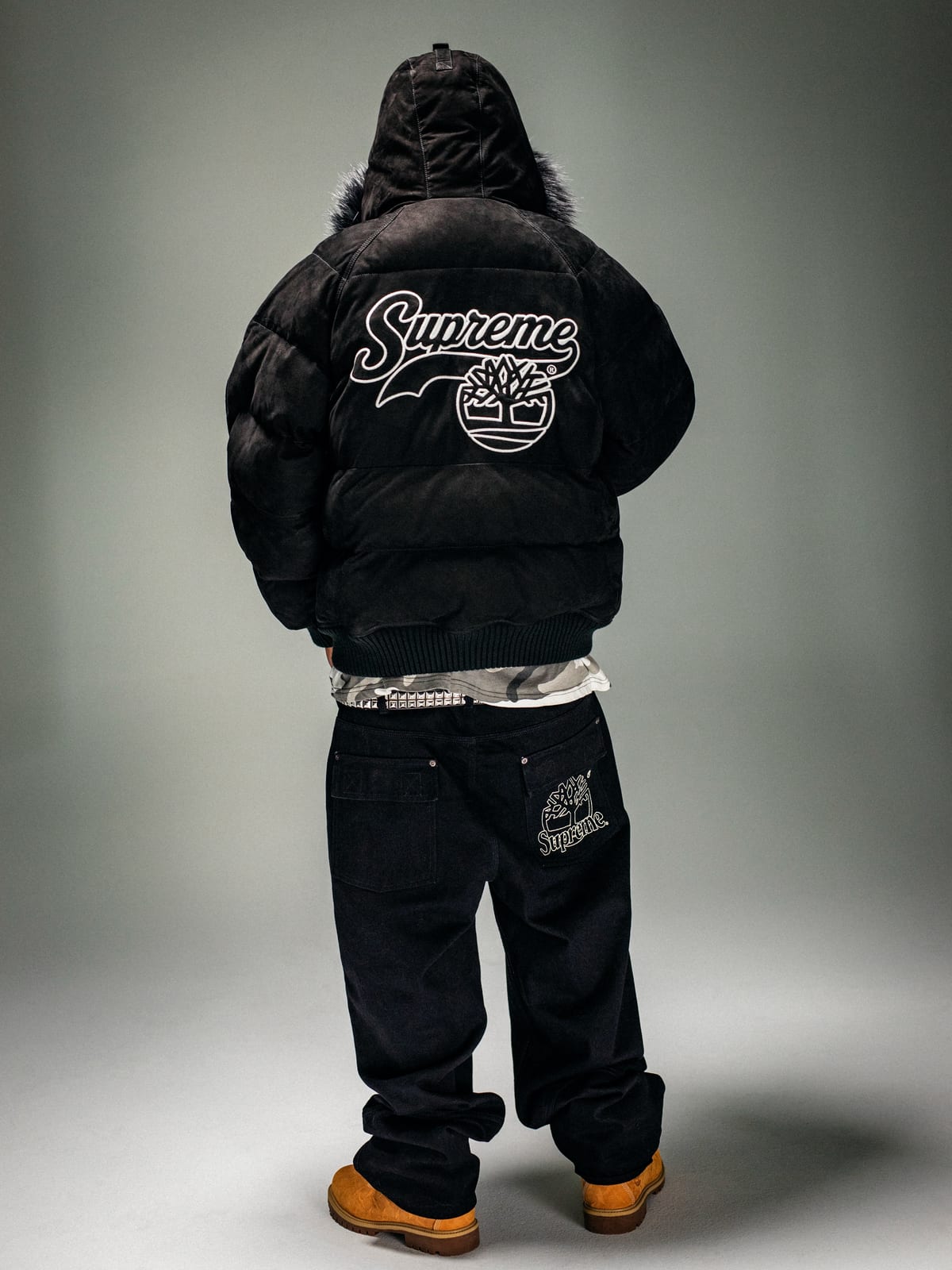 The back of a black puffer jacket with a fur-trimmed hood, printed with the Supreme script over Timberland's tree logo in a circle, worn with black jeans and wheat Timberland boots against a pale studio backdrop