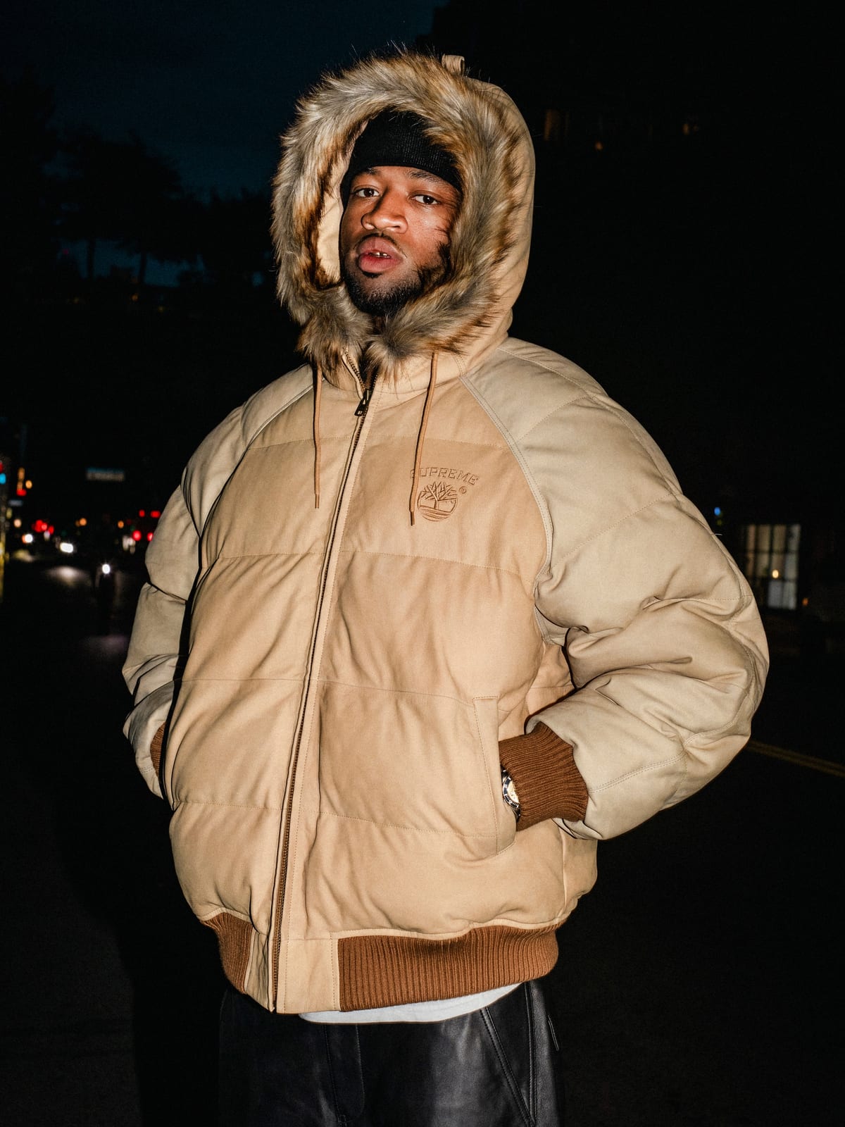 A man in a tan puffer jacket with a fur-trimmed hood and brown ribbed cuffs and hem, a small Supreme and Timberland tree logo on the chest, standing on a street at night