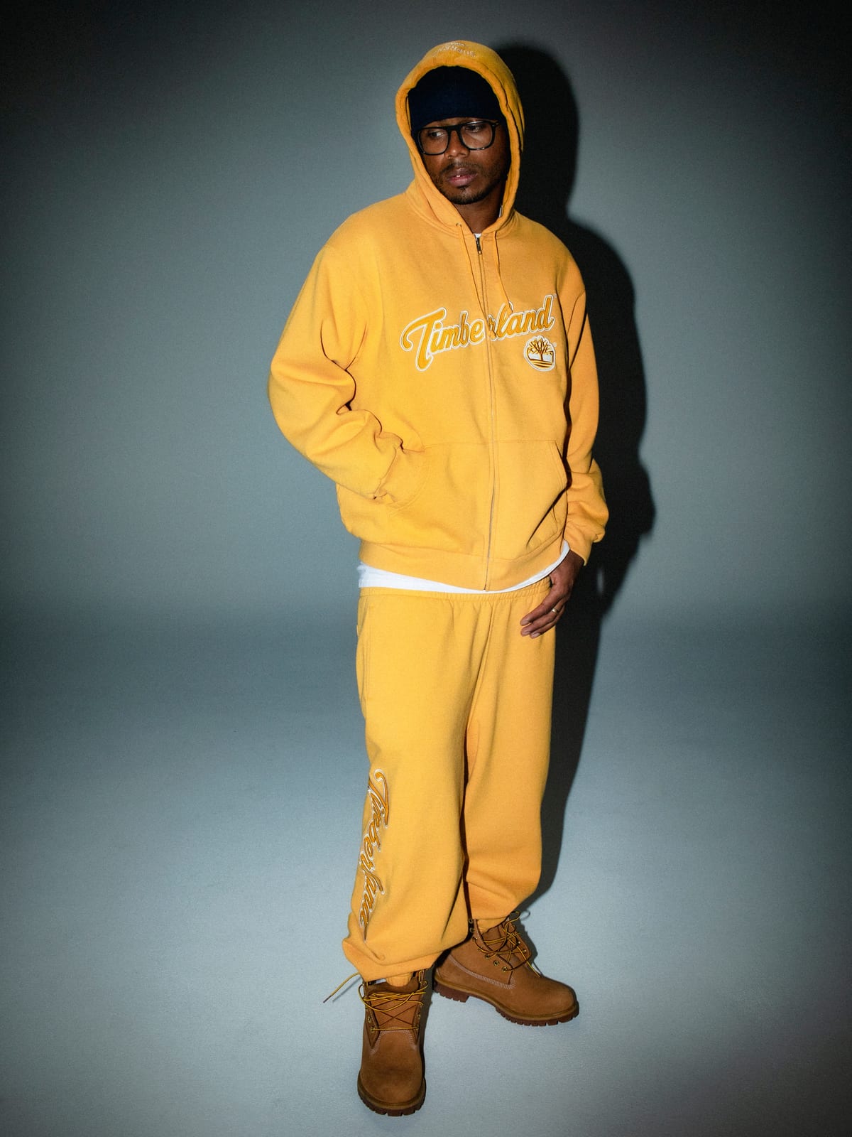 A man in a yellow zip hoodie with Timberland script lettering and matching yellow sweatpants with Timberland written down the leg, with wheat Timberland boots, against a grey studio backdrop