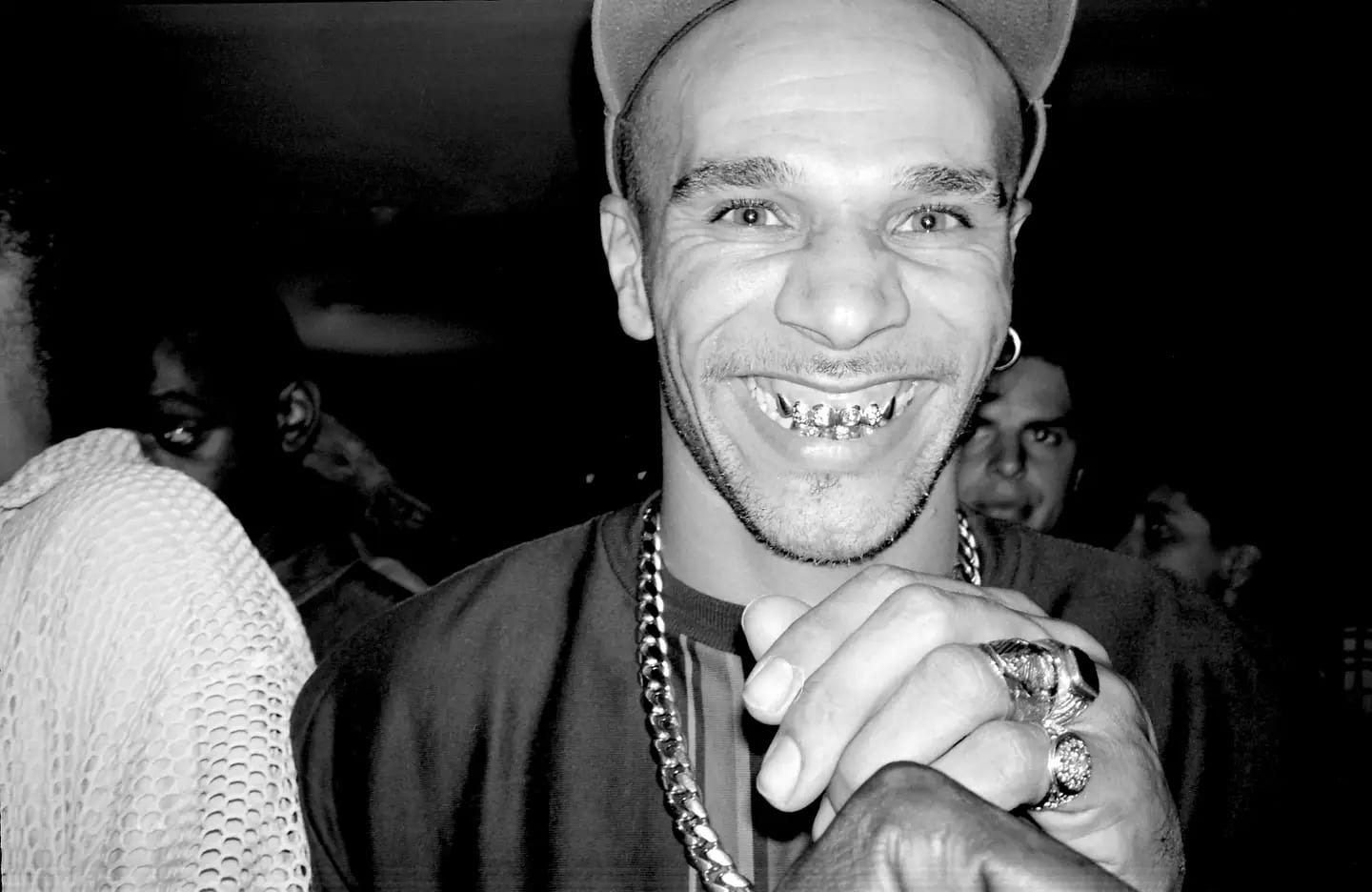 Black-and-white photograph of DJ Goldie grinning to show his gold teeth, in a cap and chain, among a crowd at the Blue Note club in London in 1996, by Normski Norman Anderson, one of the works in Tate Britain's The 90s: Art and Fashion