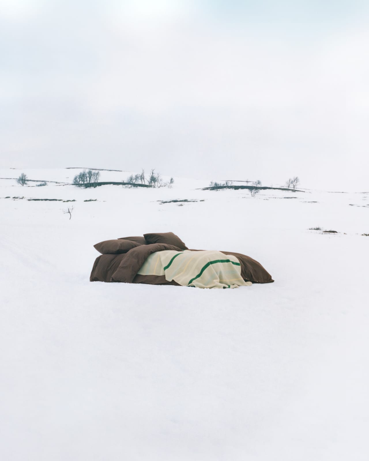 A made bed with brown pillows and duvet and a cream blanket with green stripes sits alone on a snowy plain under a pale sky, with a few bare trees in the distance