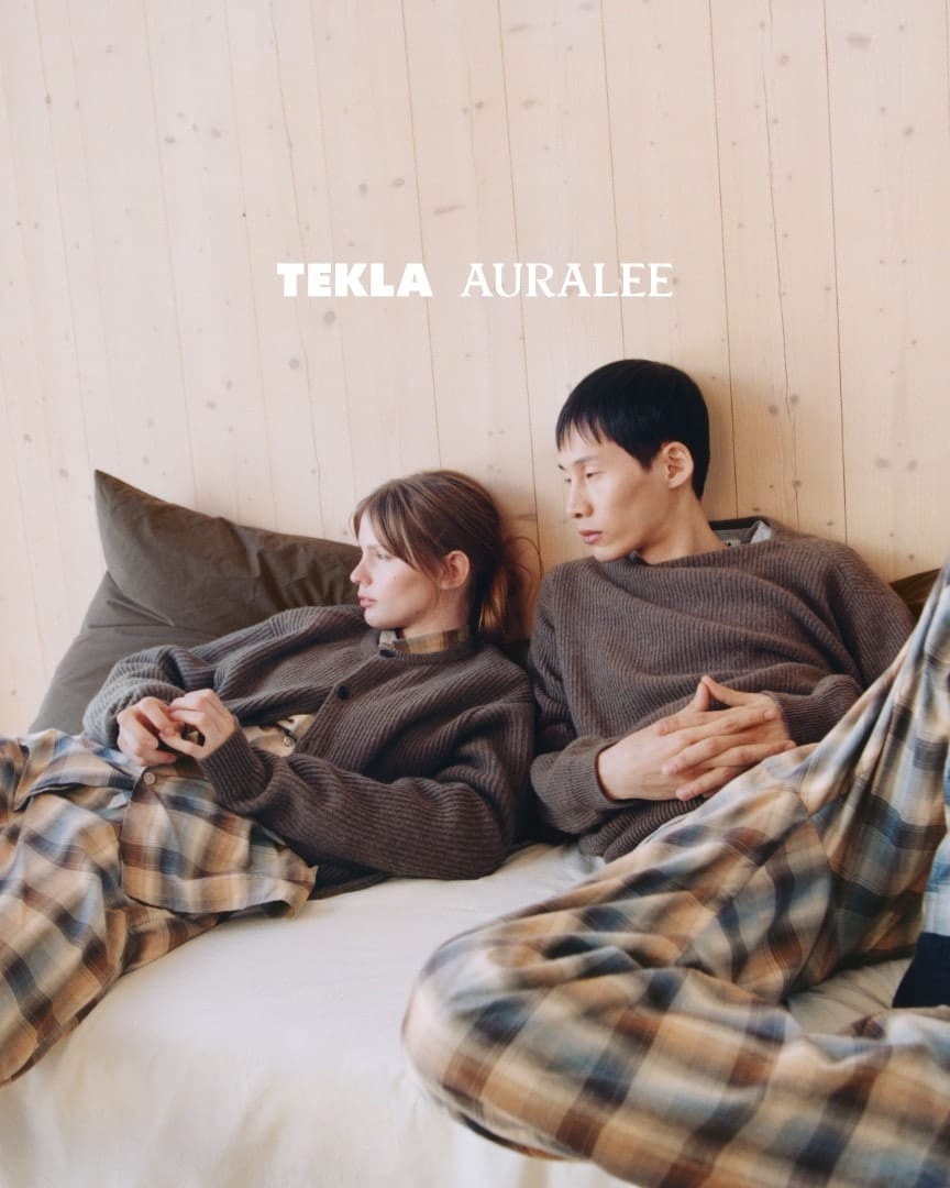 Two people sit back against pillows on a white bed by a pale wooden wall, both in brown ribbed knits over checked pyjama trousers, under the TEKLA AURALEE logo