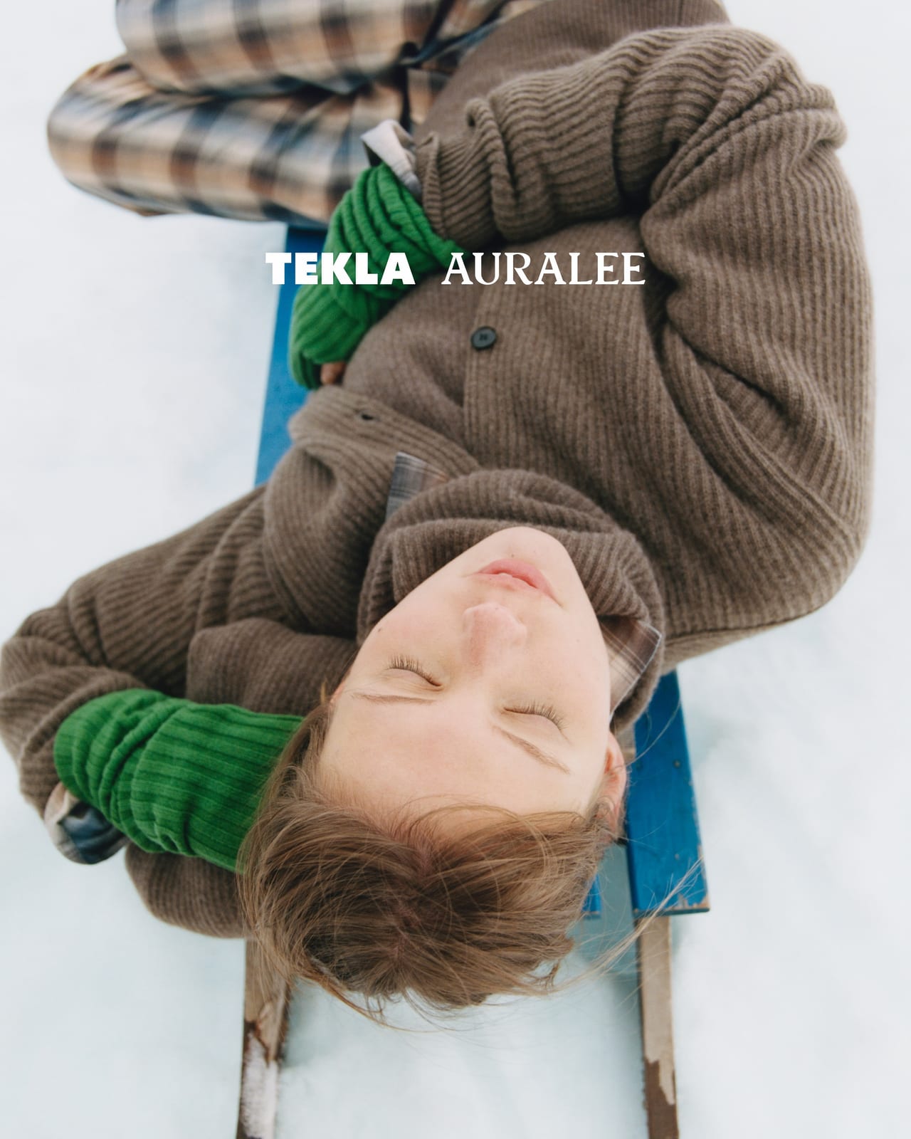 Seen from above, a person lies on a blue wooden sled in the snow, eyes closed, in a brown ribbed knit cardigan with green ribbed cuffs over checked pyjamas, under the TEKLA AURALEE logo