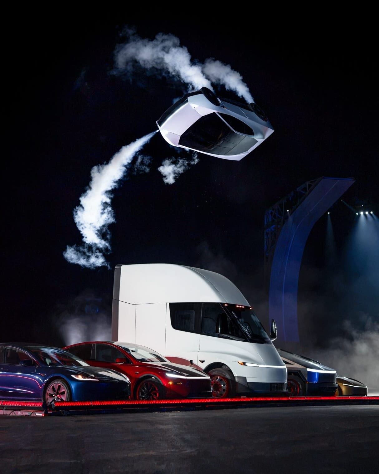 The white Tesla Roadster upside down in midair against a night sky, trailing white vapour, above a row of parked Teslas: a blue car, a red car, a white Tesla Semi and two more behind it, with a tall curved ramp lit up in blue in the background