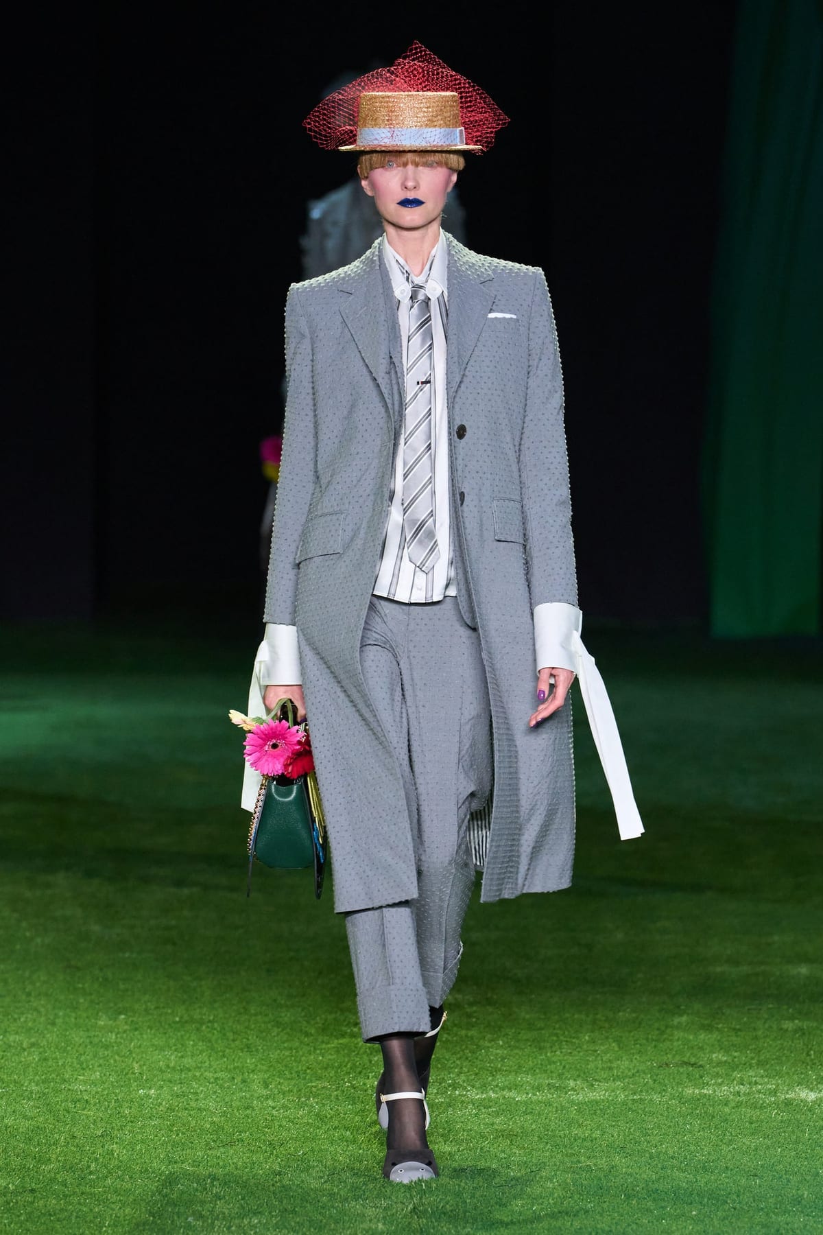 A grey tailored look from Thom Browne's Spring 2027 show: a dotted grey seersucker suit and coat with a striped tie, worn with a straw boater hat wrapped in red tulle netting