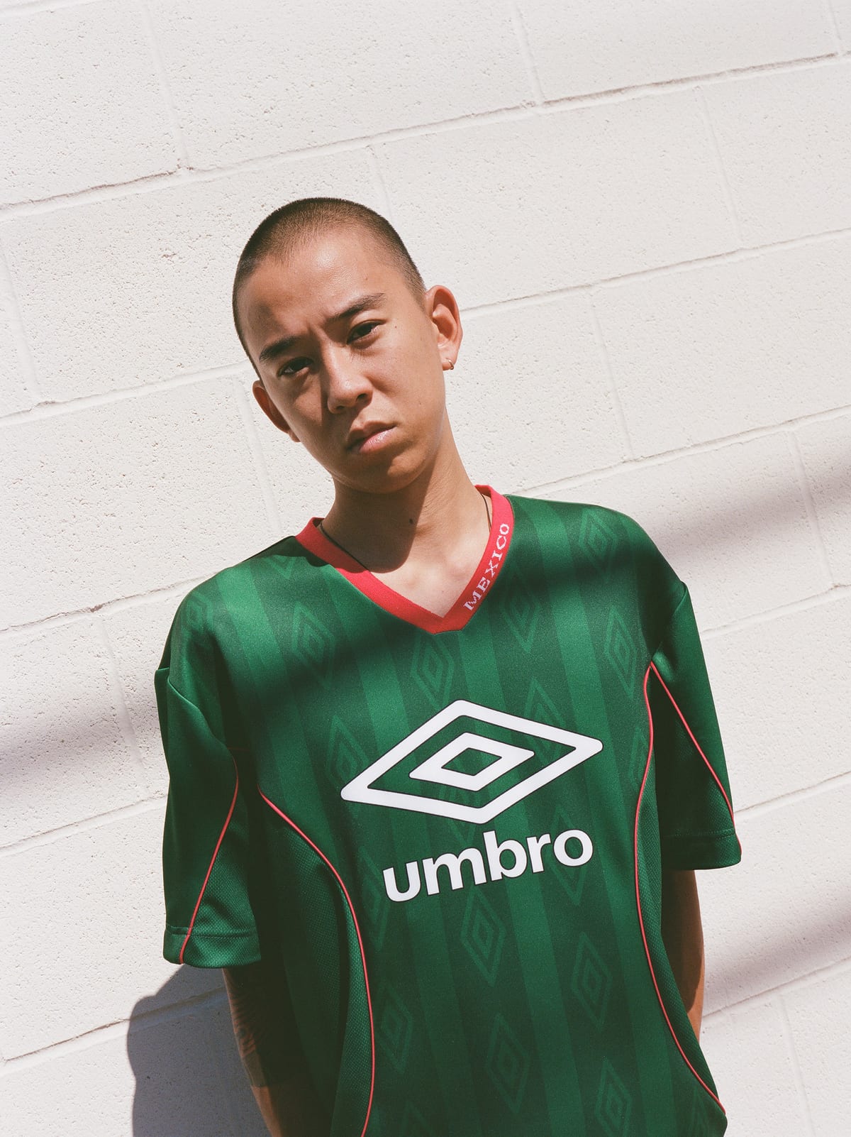 A man in a green Umbro jersey with tonal diamond stripes, red piping and a red V-neck with MEXICO printed on the collar, against a white block wall