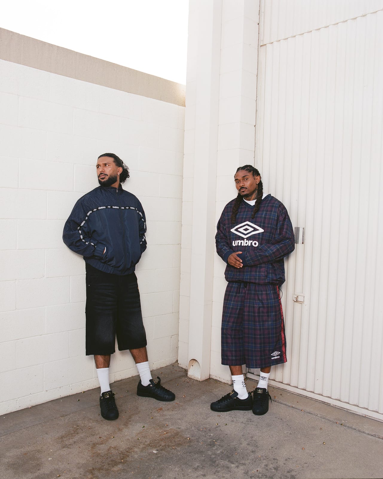Two men against a white wall: one in a navy Umbro track jacket with logo tape on the sleeves and black denim shorts, the other in a plaid Umbro pullover and matching long shorts