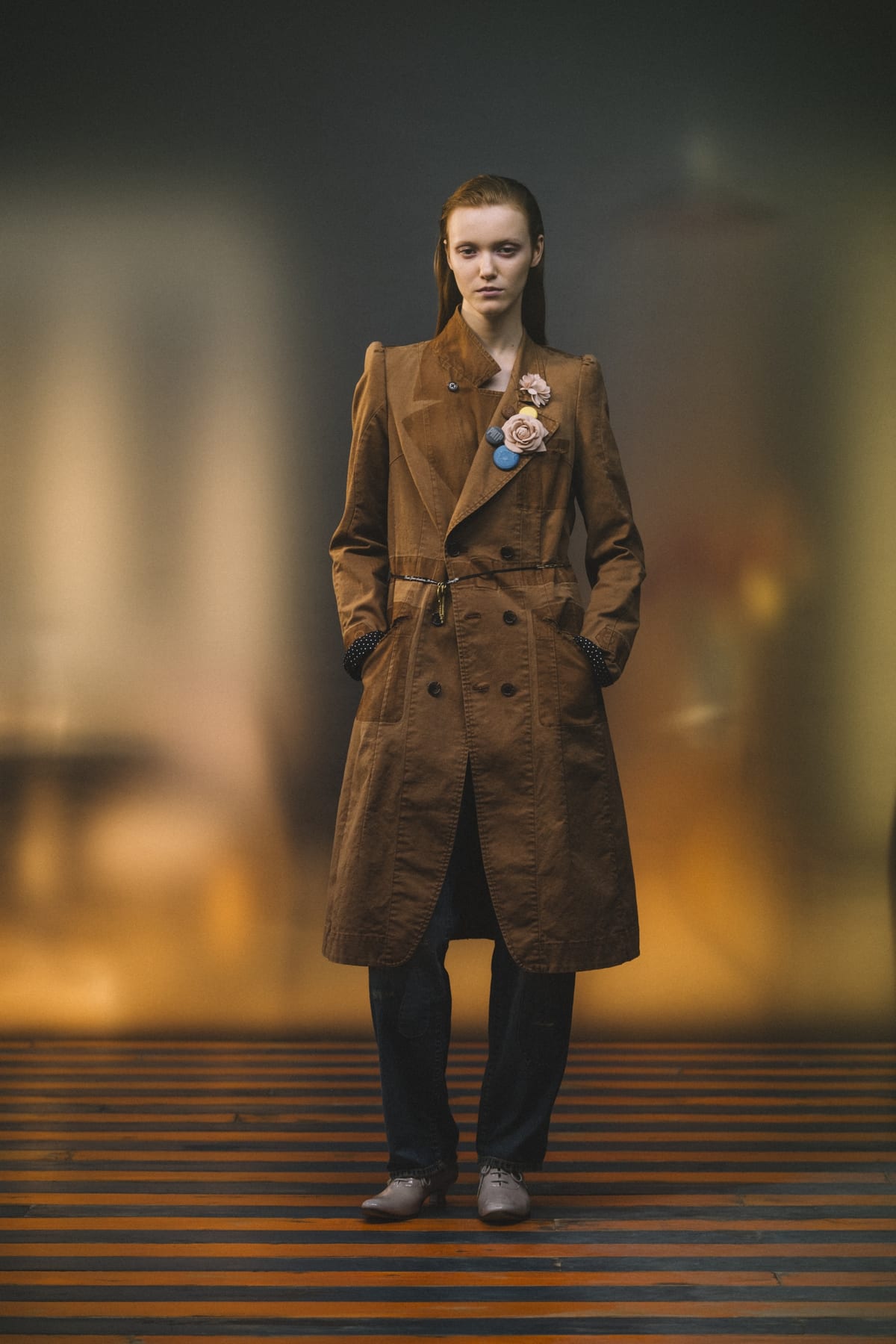 An Undercover Spring 2027 look: a sun-bleached brown belted double-breasted coat with badges and a fabric rose on the lapel, over dark trousers