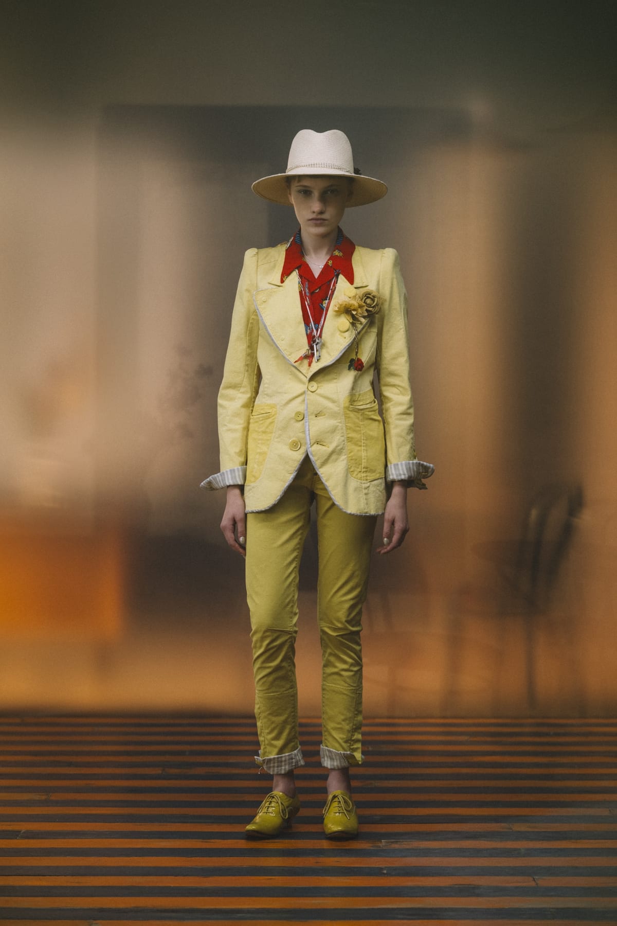 An Undercover Spring 2027 look: a pale yellow jacket with a flower pin over a red shirt, matching yellow trousers and a white hat, against a blurred orange backdrop and a striped floor