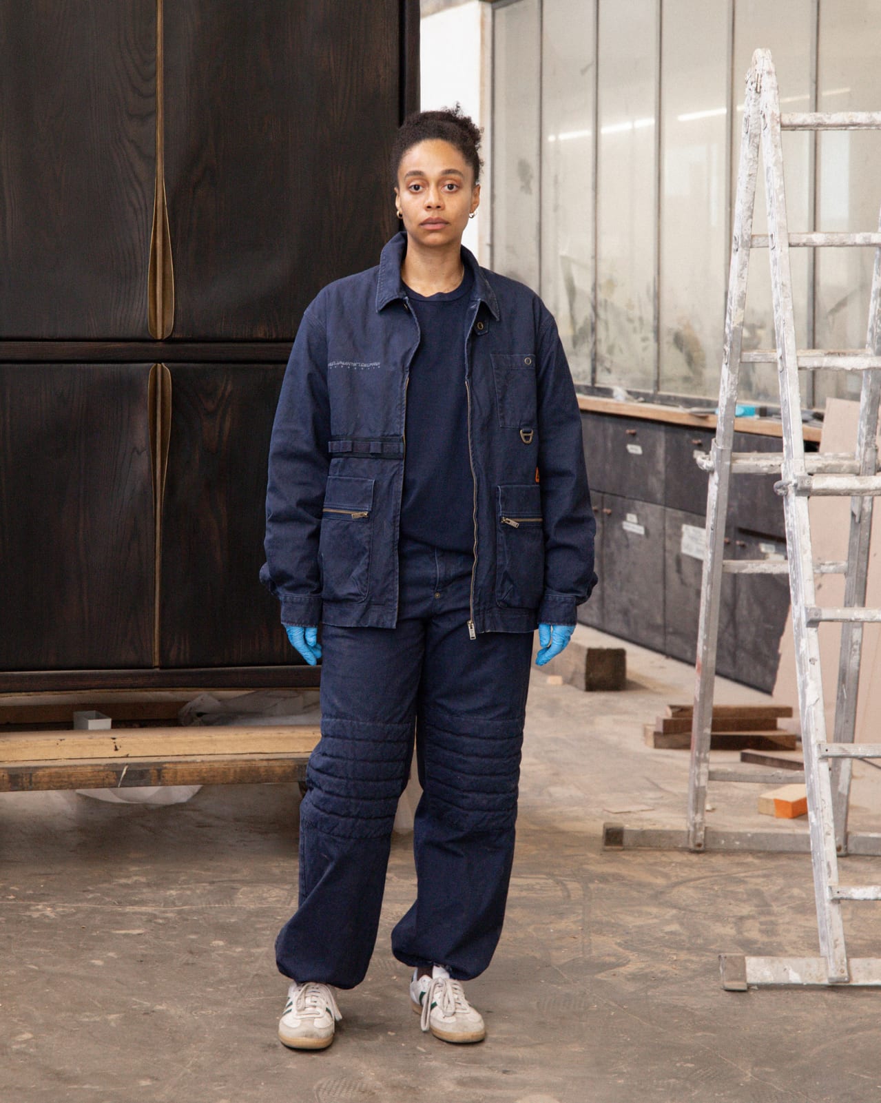 A woman in blue nitrile gloves stands on a workshop floor in a navy zip jacket and matching navy trousers with quilted knees, beside a stepladder and a tall dark wooden cabinet