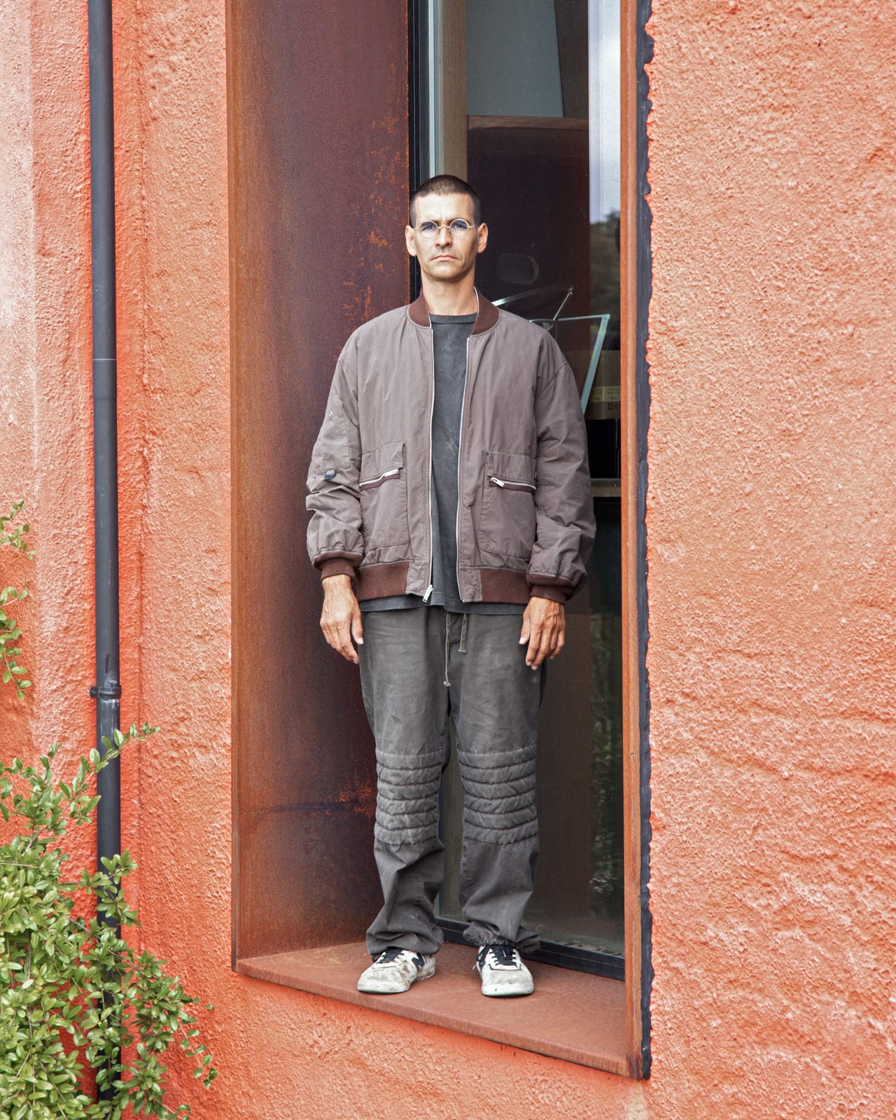 A man in glasses stands in a doorway set into a rust-red rendered wall, in a taupe bomber jacket with a brown ribbed collar over a dark T-shirt, and grey trousers with quilted knee panels