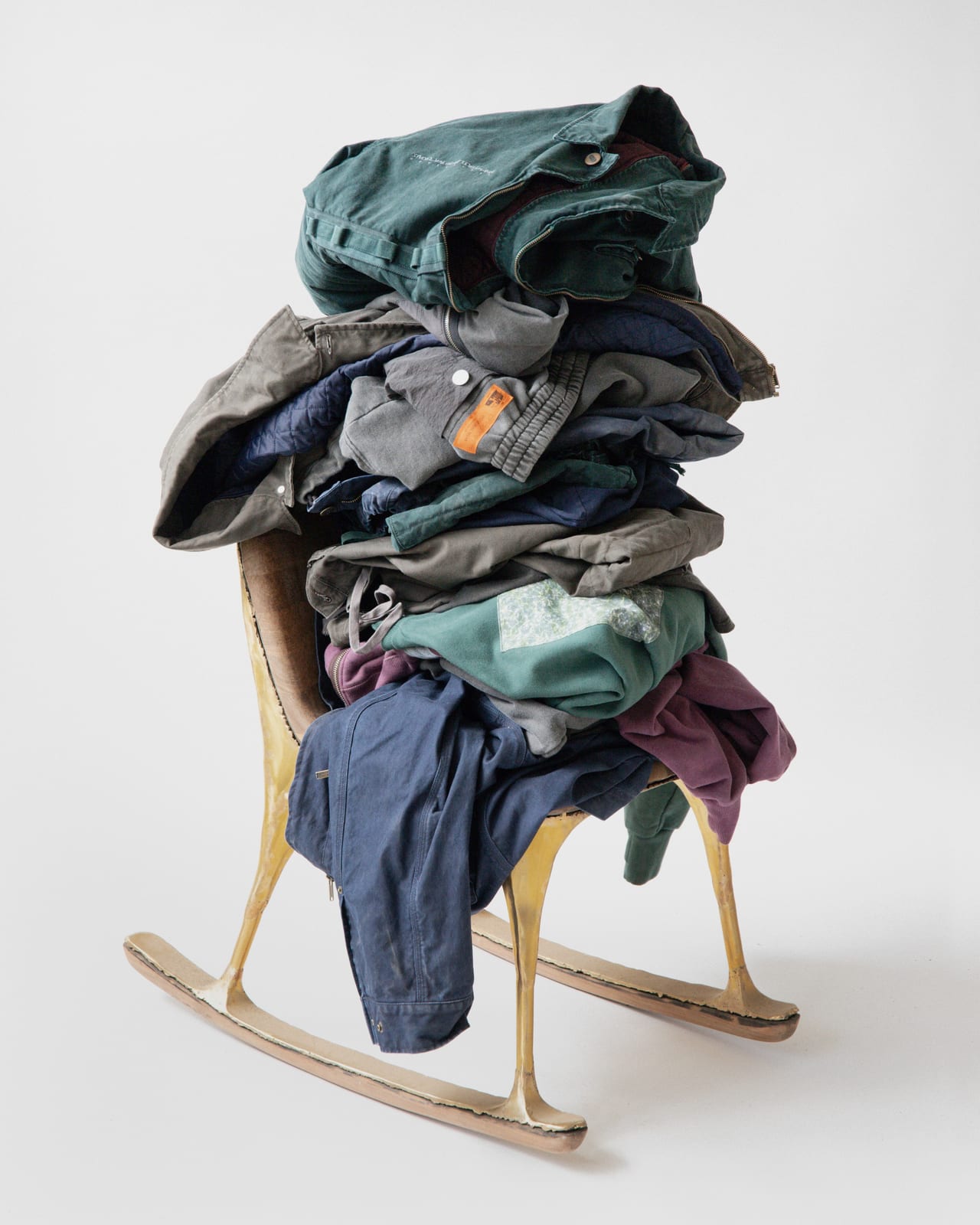 A tall pile of folded and crumpled workwear in green, grey, navy and plum, heaped on a wooden rocking chair with brass-coloured legs, against a white backdrop