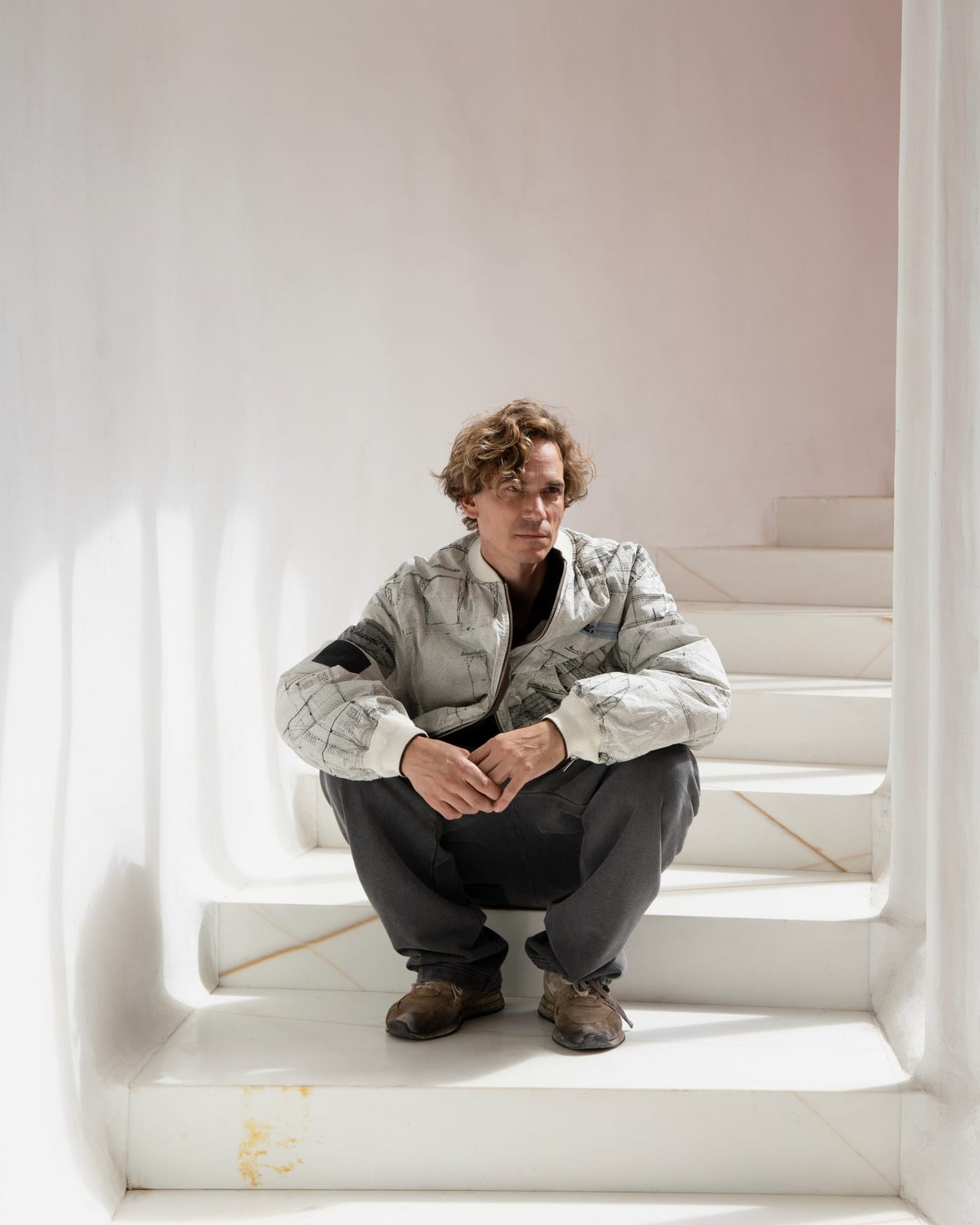 A man with curly hair crouches on white plastered stairs in an off-white bomber jacket printed all over with black line drawings, with cream ribbed trim, and charcoal trousers