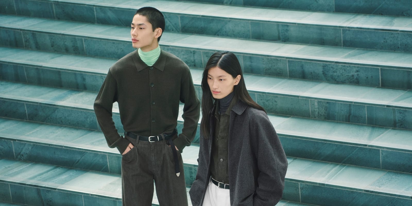 Two models on teal stone steps in the UNIQLO : C Autumn/Winter 2026 campaign designed by Clare Waight Keller, one in an olive ribbed knit shirt over a mint turtleneck, the other in a charcoal wool jacket over an olive shirt with white trousers