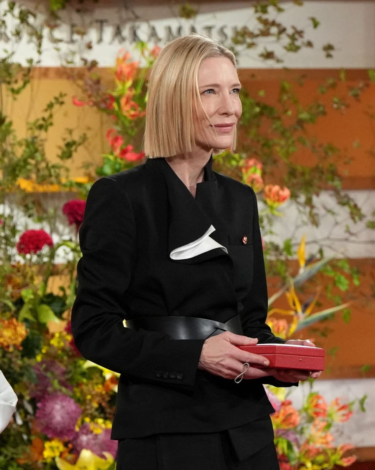 Cate Blanchett, with a blonde bob, in a black suit jacket with a sculpted collar, a white silk fold at the lapel and a wide black leather belt, holding a red presentation box in front of a wall of orange and red flowers