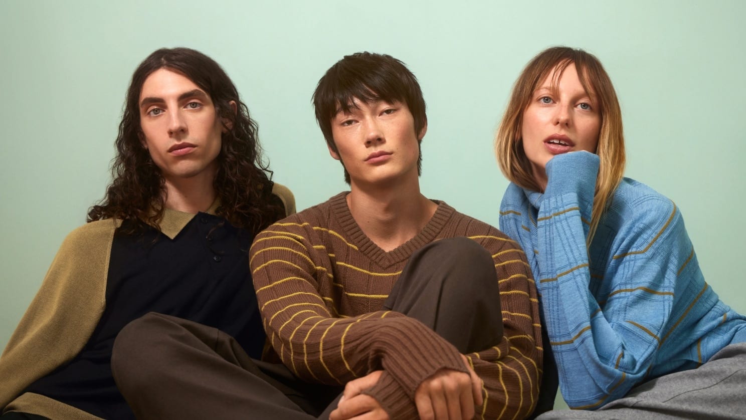 Three models sitting against a pale mint wall in UNIQLO F.RISSO knits: on the left a long-haired man in a navy polo sweater with khaki sleeves and collar, in the middle a man in a brown V-neck with thin yellow stripes, and on the right a woman in a light blue turtleneck with brown stripes