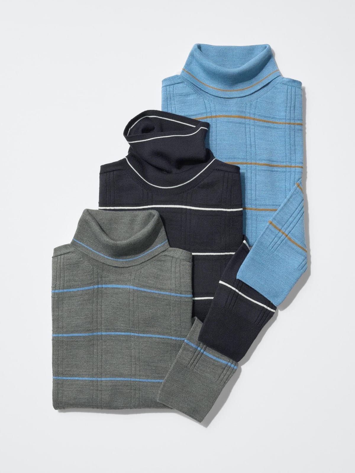 Three UNIQLO F.RISSO 3D knit turtleneck sweaters laid out overlapping on a white background: light blue with mustard stripes at the back, navy with white stripes in the middle and grey with blue stripes in front