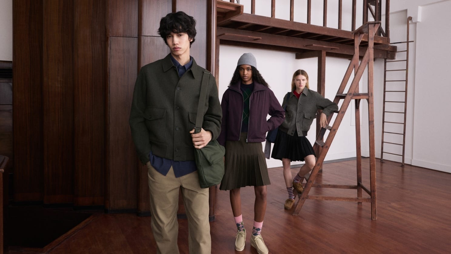 Three models in a wood-panelled studio with a wooden ladder and gallery: a young man in a dark green wool shirt jacket, khaki trousers and a green shoulder bag, a woman in a plum zip blouson, grey beanie, argyle sweater and olive pleated skirt, and a woman in a grey check blouson and black pleated skirt leaning on the ladder
