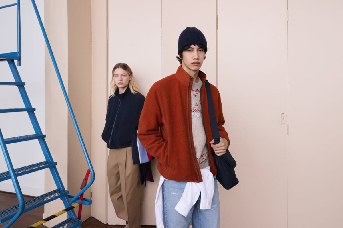A young man in a navy beanie, rust-orange fleece zip jacket over a Fair Isle sweater, a white shirt tied at the waist and light jeans, with a navy shoulder bag, beside a woman in a navy fleece jacket and khaki trousers next to a blue metal staircase