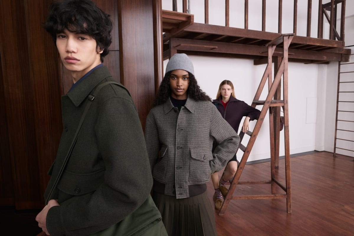 A young man in a dark green wool shirt jacket with a shoulder strap across it, a woman behind him in a grey houndstooth blouson, grey beanie and olive pleated skirt, and a third model in a plum zip jacket leaning on a wooden ladder
