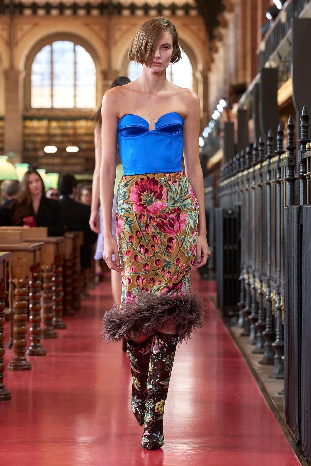 A Valentino Spring 2027 look: a strapless royal-blue satin corset top over a floral-print skirt with a dark feathered hem and floral boots, on a red runway between the library's reading tables