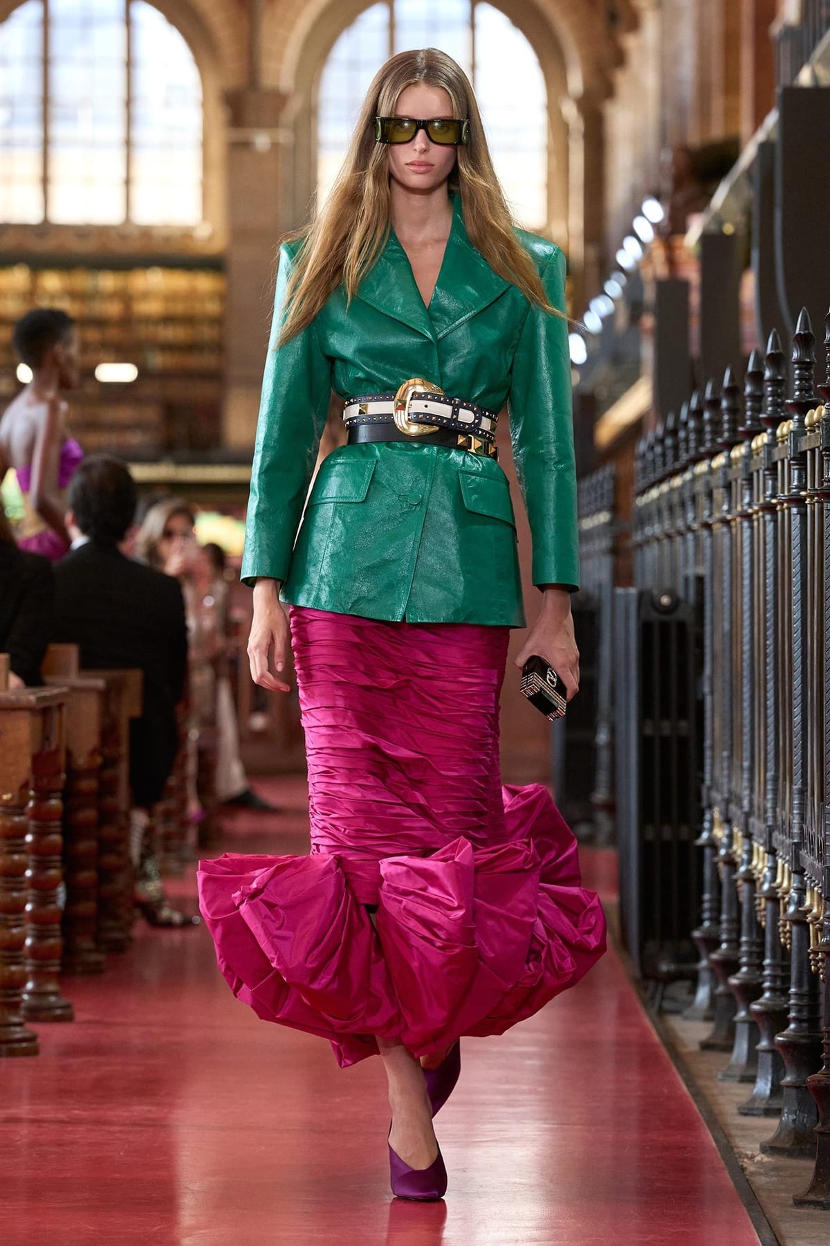 A Valentino Spring 2027 look: a belted green leather jacket over a ruched magenta skirt with a flounced hem, with tinted sunglasses and a small bag, in the library's reading room