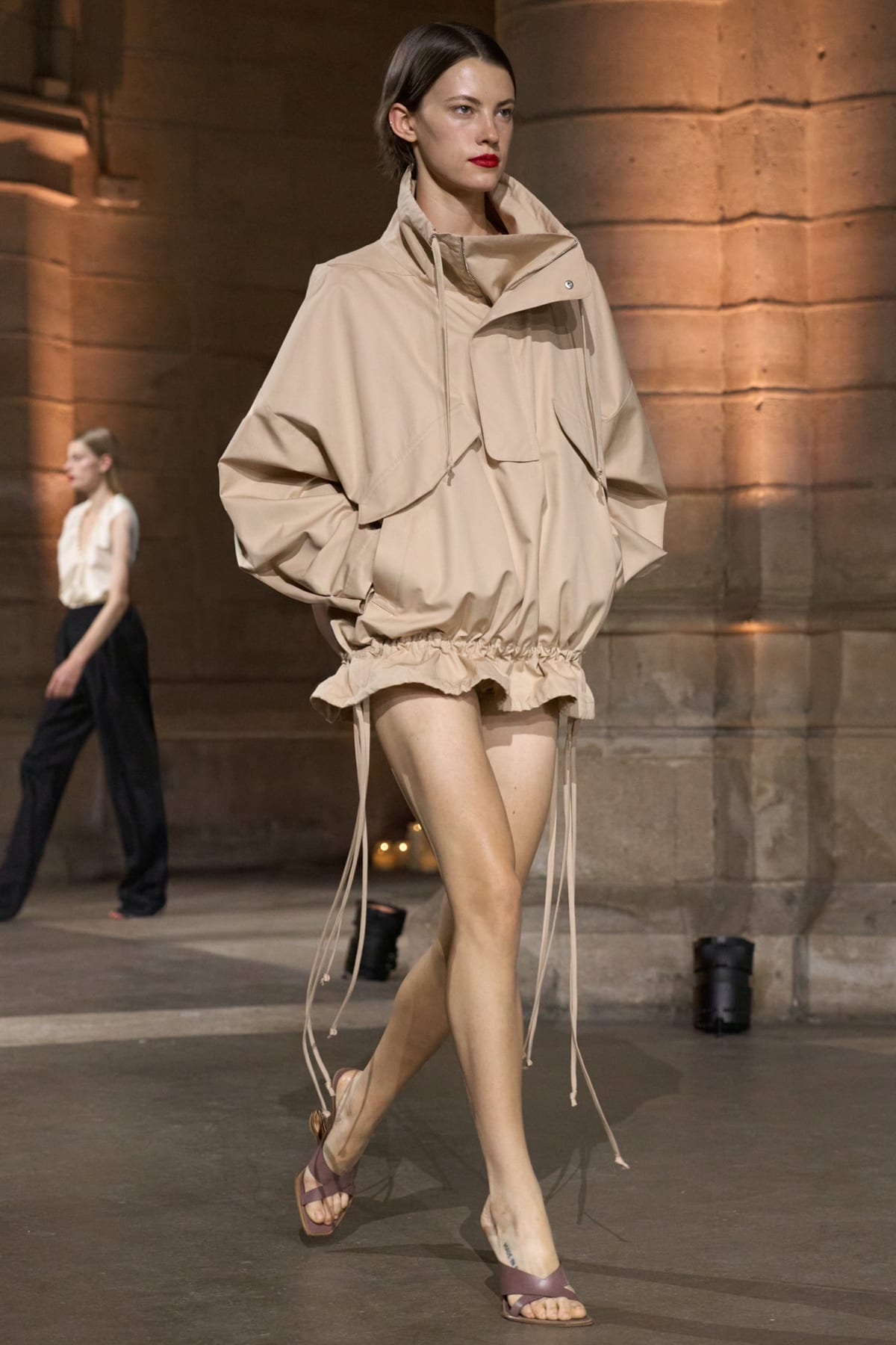 A Victoria Beckham Spring 2027 look: an oversized beige hooded anorak with a drawstring hem worn as a mini dress, with brown leather thong sandals, in the stone hall of the Conciergerie