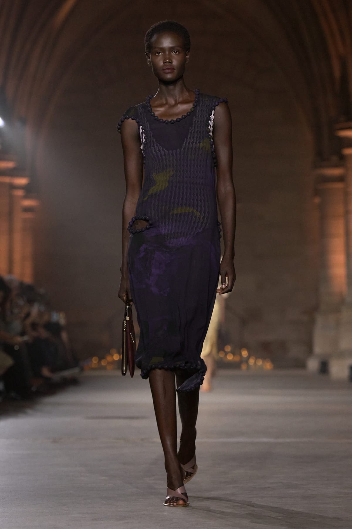 A Victoria Beckham Spring 2027 look: a sleeveless dark purple knit dress with a scalloped neckline, a torn-edge cutout at the hip and a ruffled hem, with a small brown bag and metallic sandals