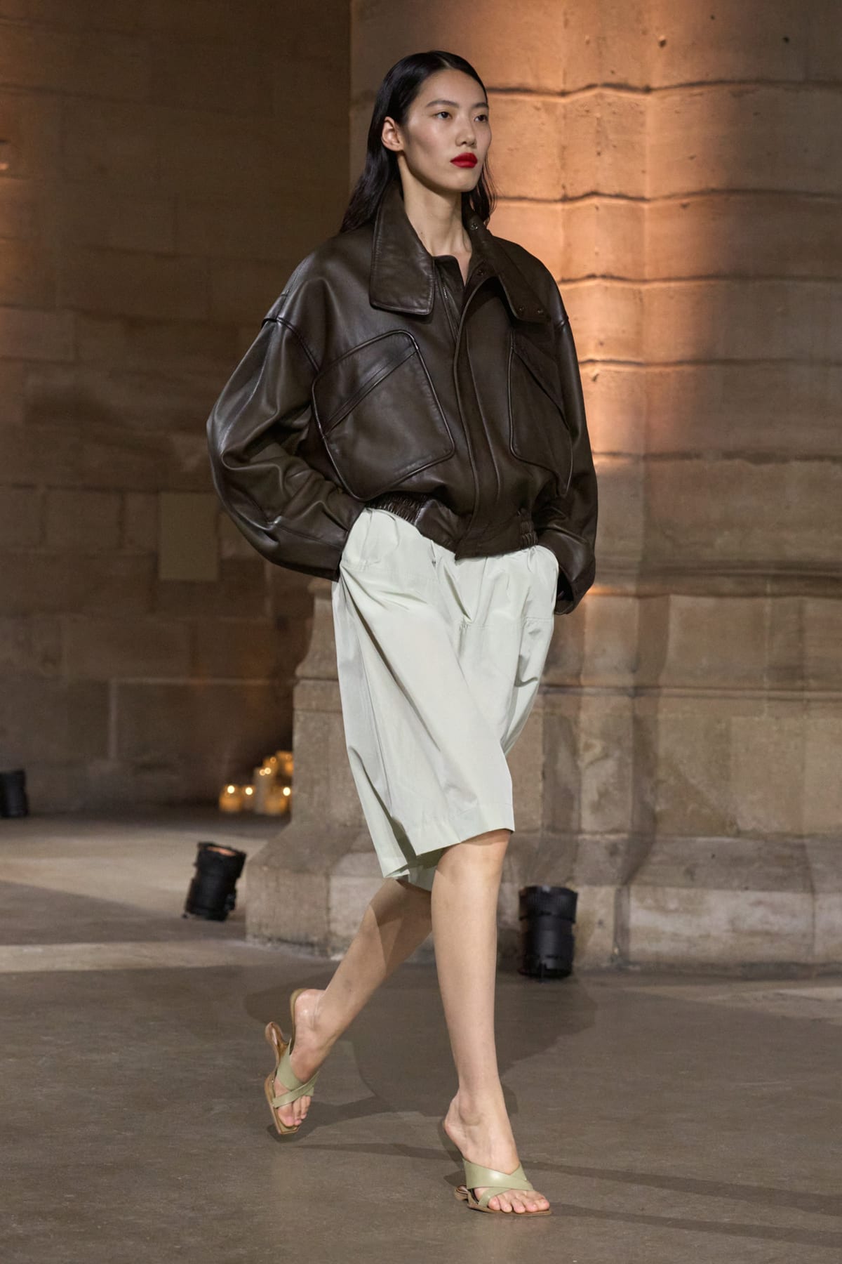 A Victoria Beckham Spring 2027 look: a chocolate-brown leather blouson jacket with a rounded shoulder over pale mint knee-length shorts, with olive thong sandals